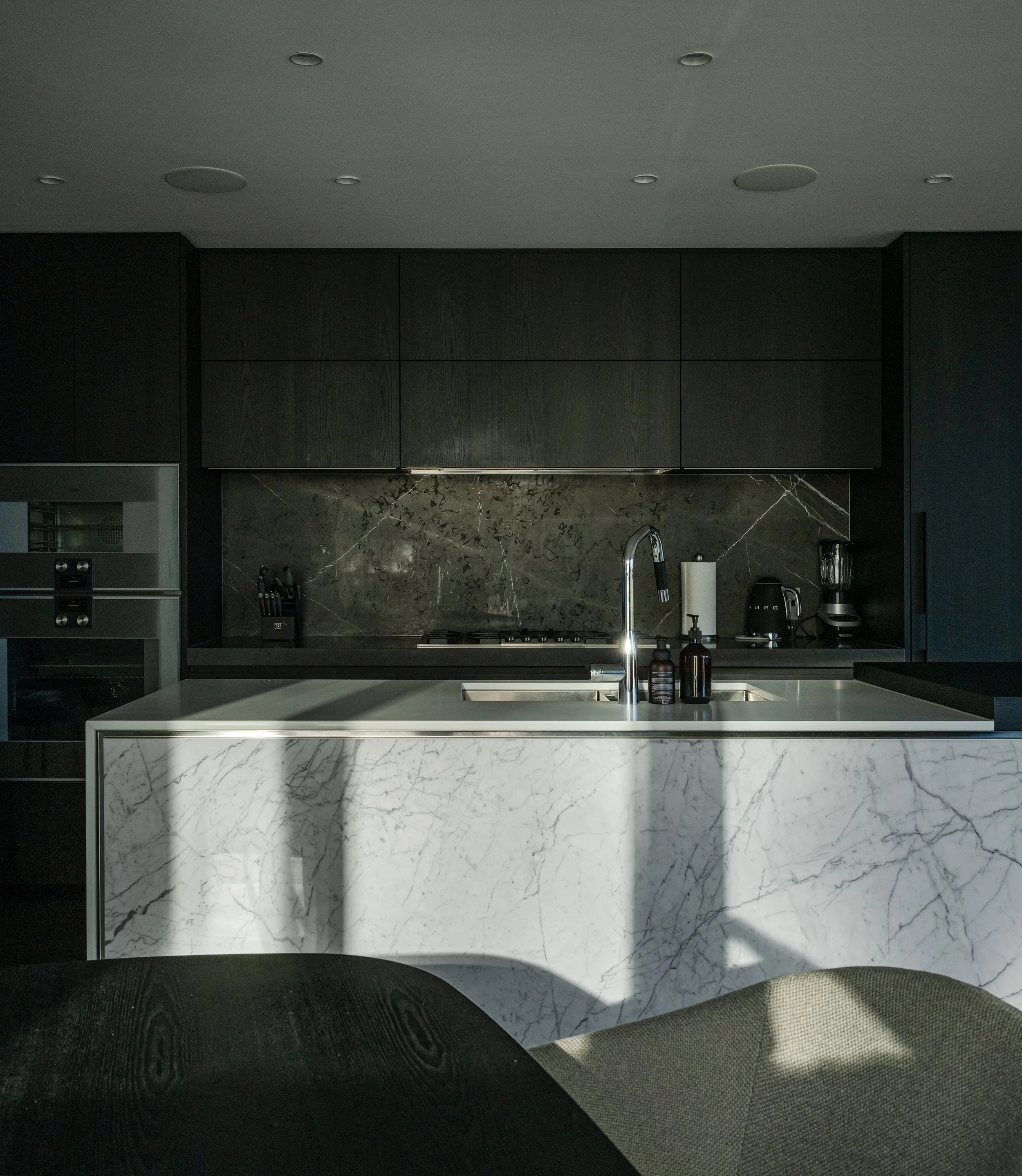 Modern kitchen with a white marble island, dark cabinets, and a built-in oven. Recessed ceiling lights and a dark wooden floor.