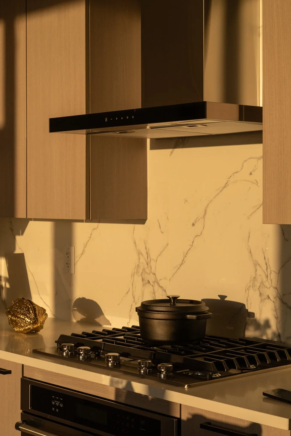 Modern kitchen with light wood cabinets, white marble backsplash, stainless steel appliances, a white island with a light wood base, large windows with city views, and gray wood flooring.
