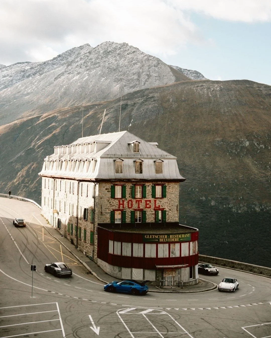 A multi-story hotel building situated on a curving mountain road with parked cars, mountain landscape background, and a cloudy sky.