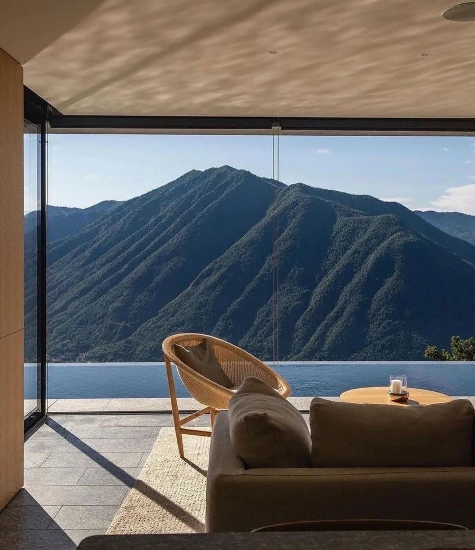 Living room with a sofa, wooden chair, round table with candle, and large glass window showing mountains and water outside.