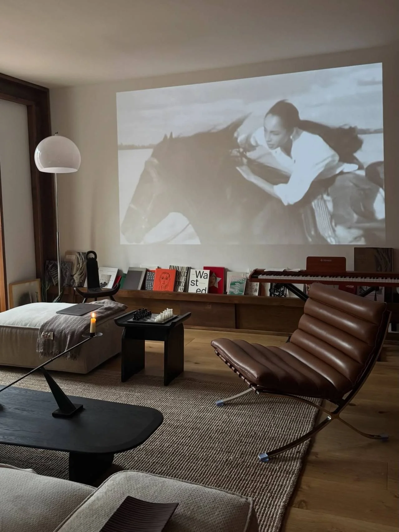 Living room with a wall-mounted black-and-white movie projection of a woman riding a horse. The room features a brown leather lounge chair, a beige cushioned chair, a small black table with a candle and chess set, a floor lamp, a wooden bookshelf wit