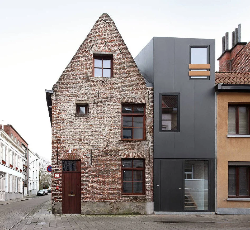 A brick house with a pitched roof next to a modern gray building with flat roof on a city street.