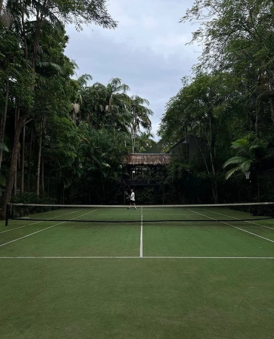 Tennis court surrounded by dense trees with a person playing tennis in the background, overcast sky.