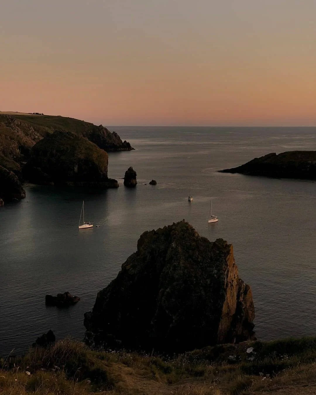 Seaside view during sunset with large rocks and cliffs, calm water, and sailboats anchored near the shore.