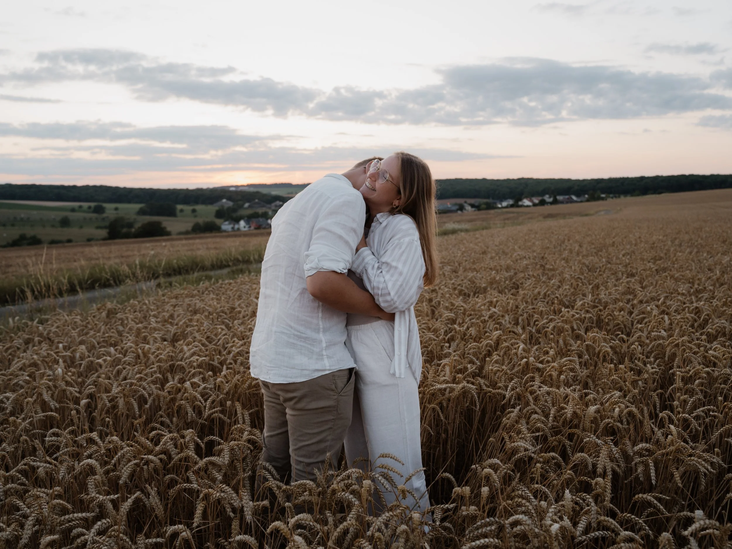 A couple hugging and smiling in a wheat field during sunset, with a rural landscape and sky with clouds in the background.
