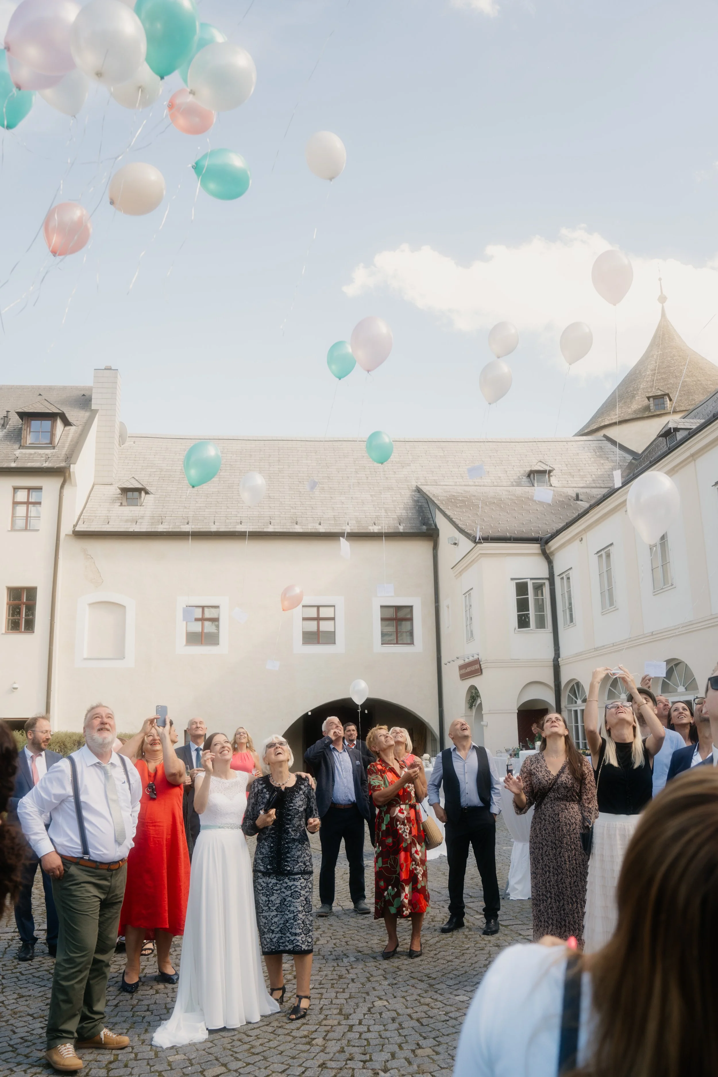 People releasing pastel-colored balloons outdoors in a courtyard during a celebration or event.