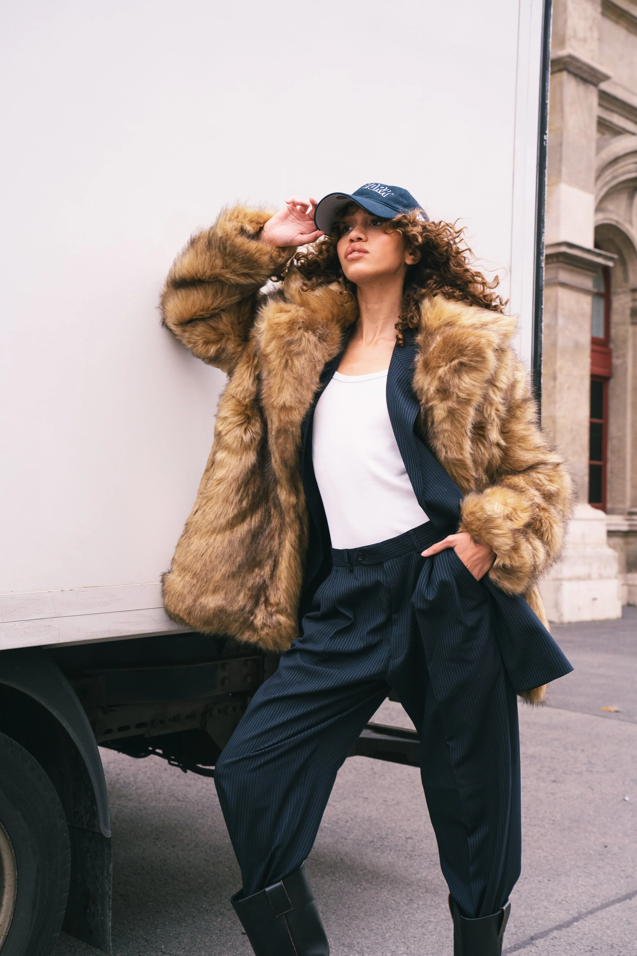 Woman wearing a fur coat, white top, pinstripe pants, black boots, and a cap, standing against a white wall on a city street.