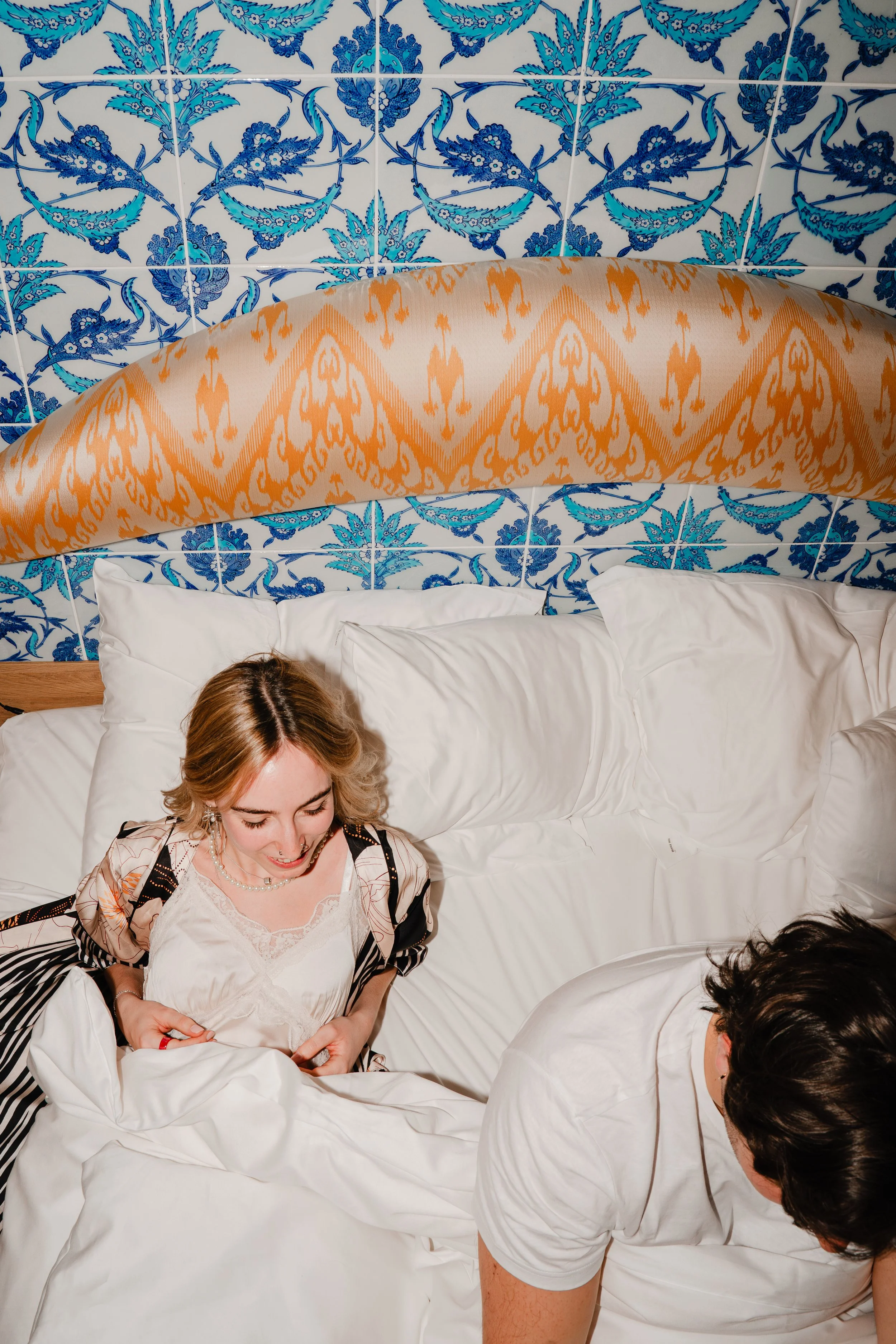 Two people sitting on a bed, with a blue and white floral tiled wall in the background. One person with light brown hair is smiling, wearing a lace-trimmed top with a patterned robe. The other person has dark hair and is leaning forward with their he