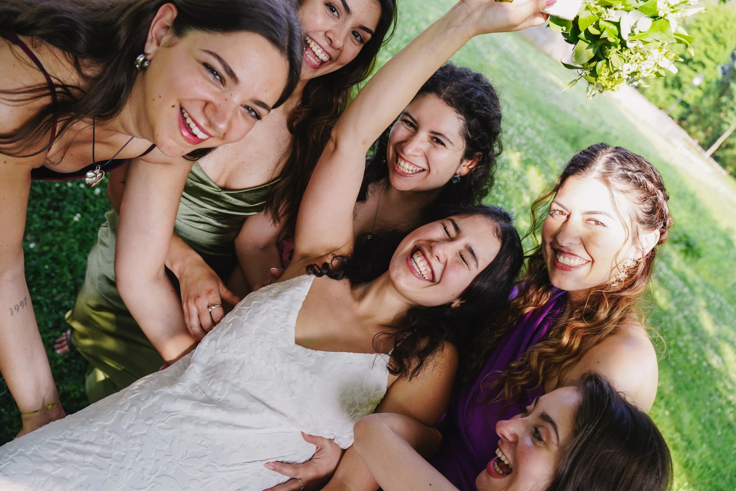 Group of women outdoors smiling, one wearing a white dress, others in colorful dresses, on green grass with trees in the background.