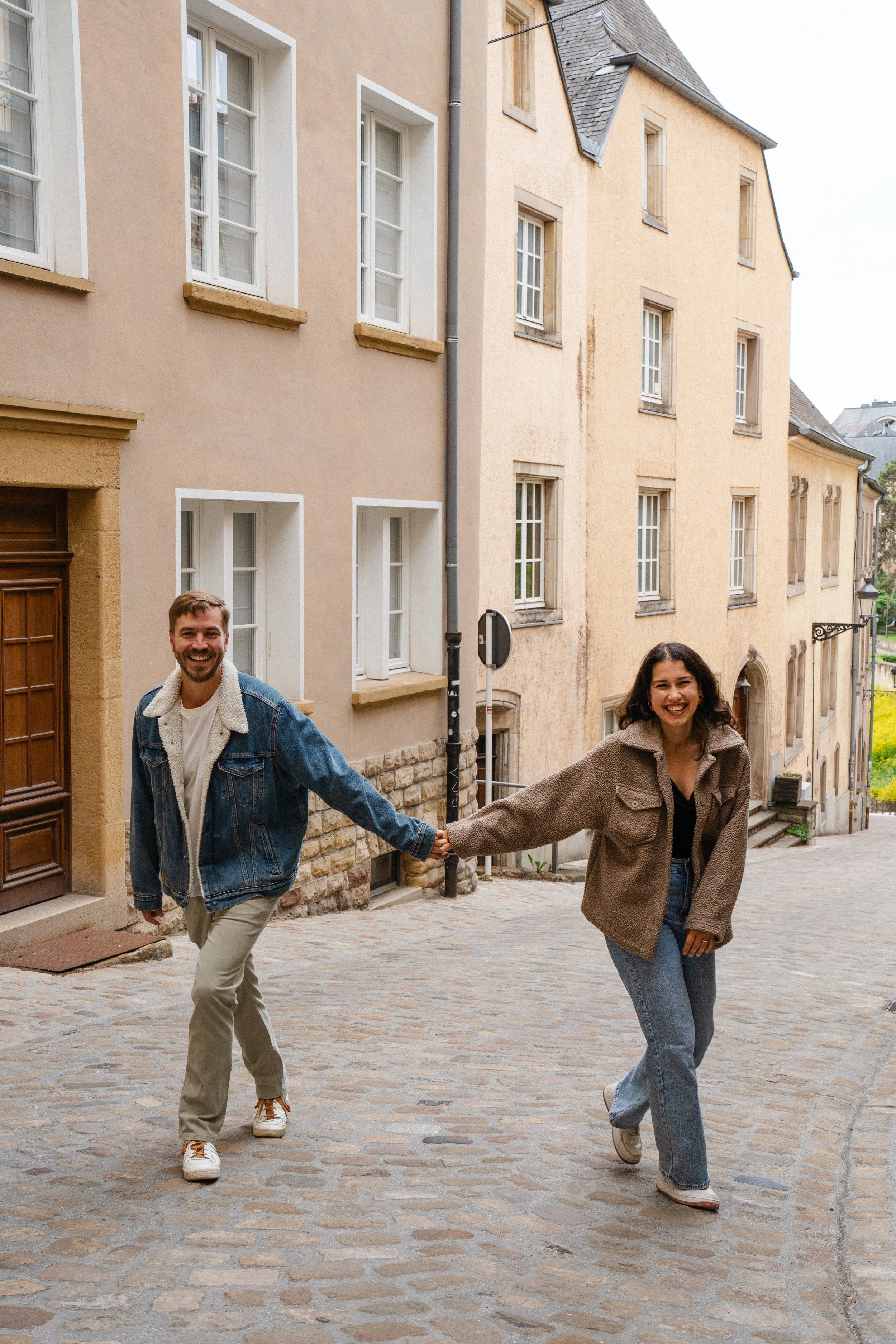 A man and woman are holding hands and smiling while walking down a cobblestone street in a European city, with colorful old buildings in the background.