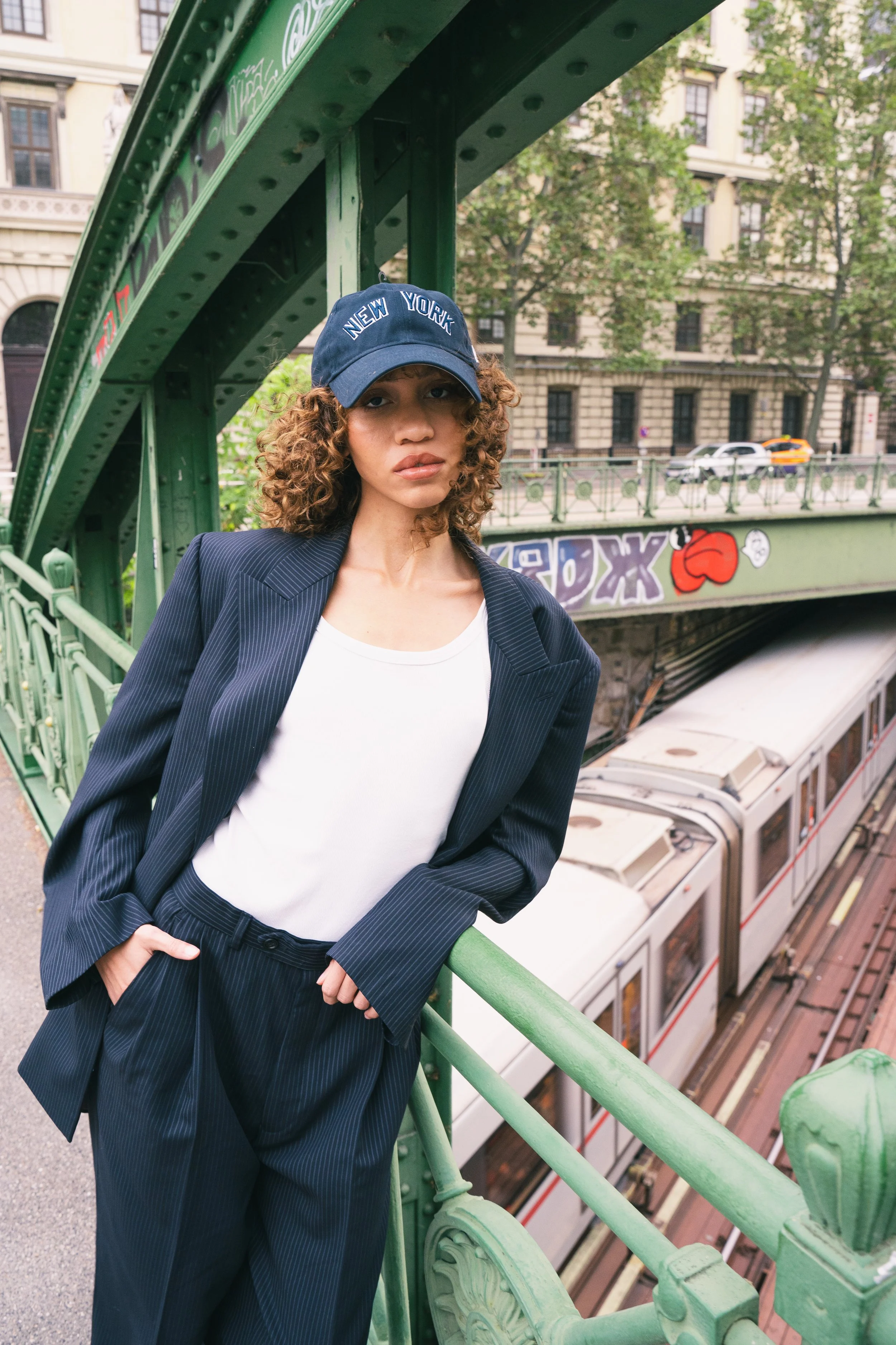 Young woman with curly hair wearing a dark blue New York cap, a pinstripe blazer, and a white t-shirt, leaning on a green railing of an elevated train station platform in New York City, with a train below and graffiti on the wall behind her.