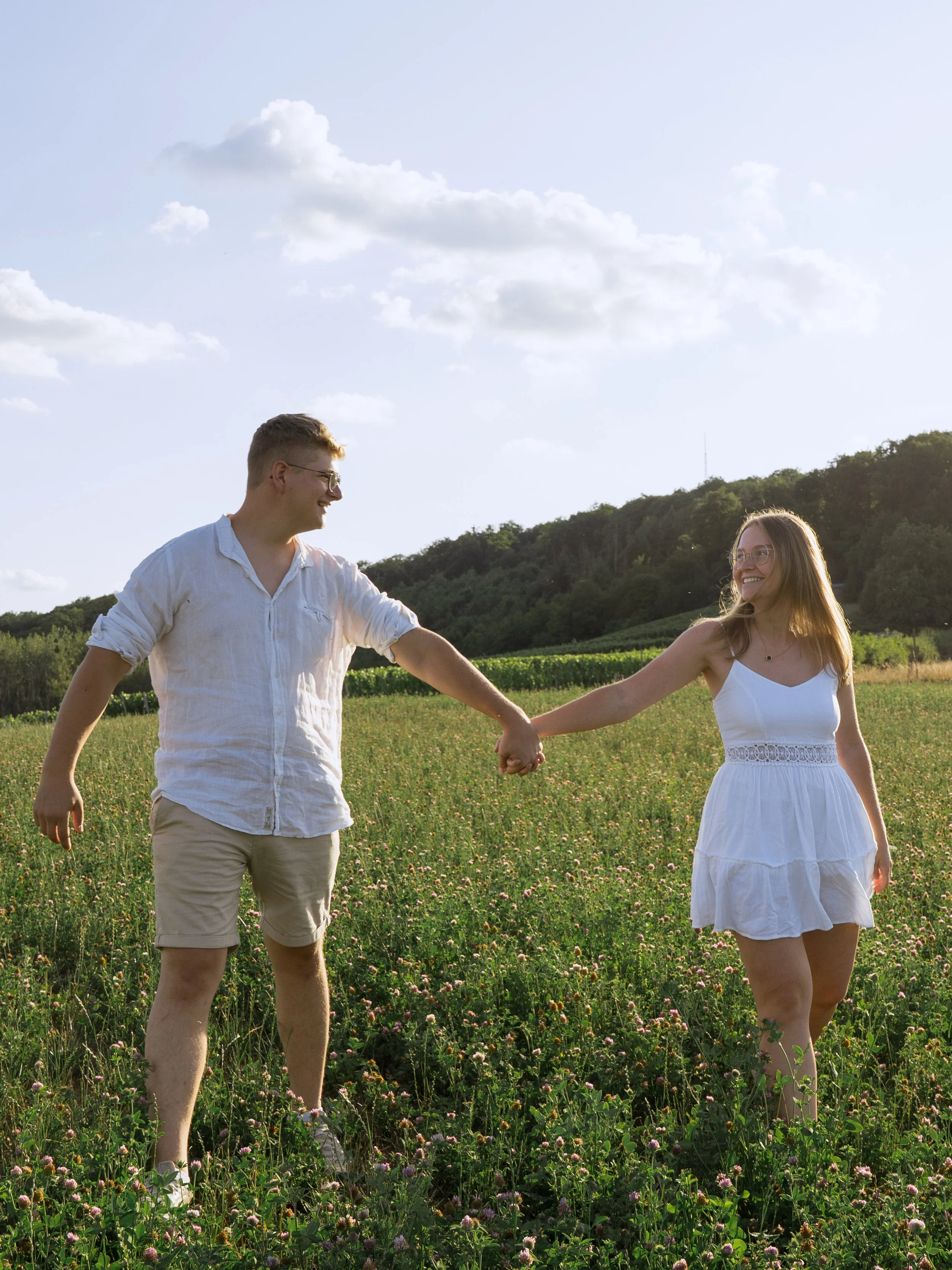 A young couple holding hands and smiling while walking through a field of green grass and pink flowers, with a hillside and blue sky with clouds in the background.