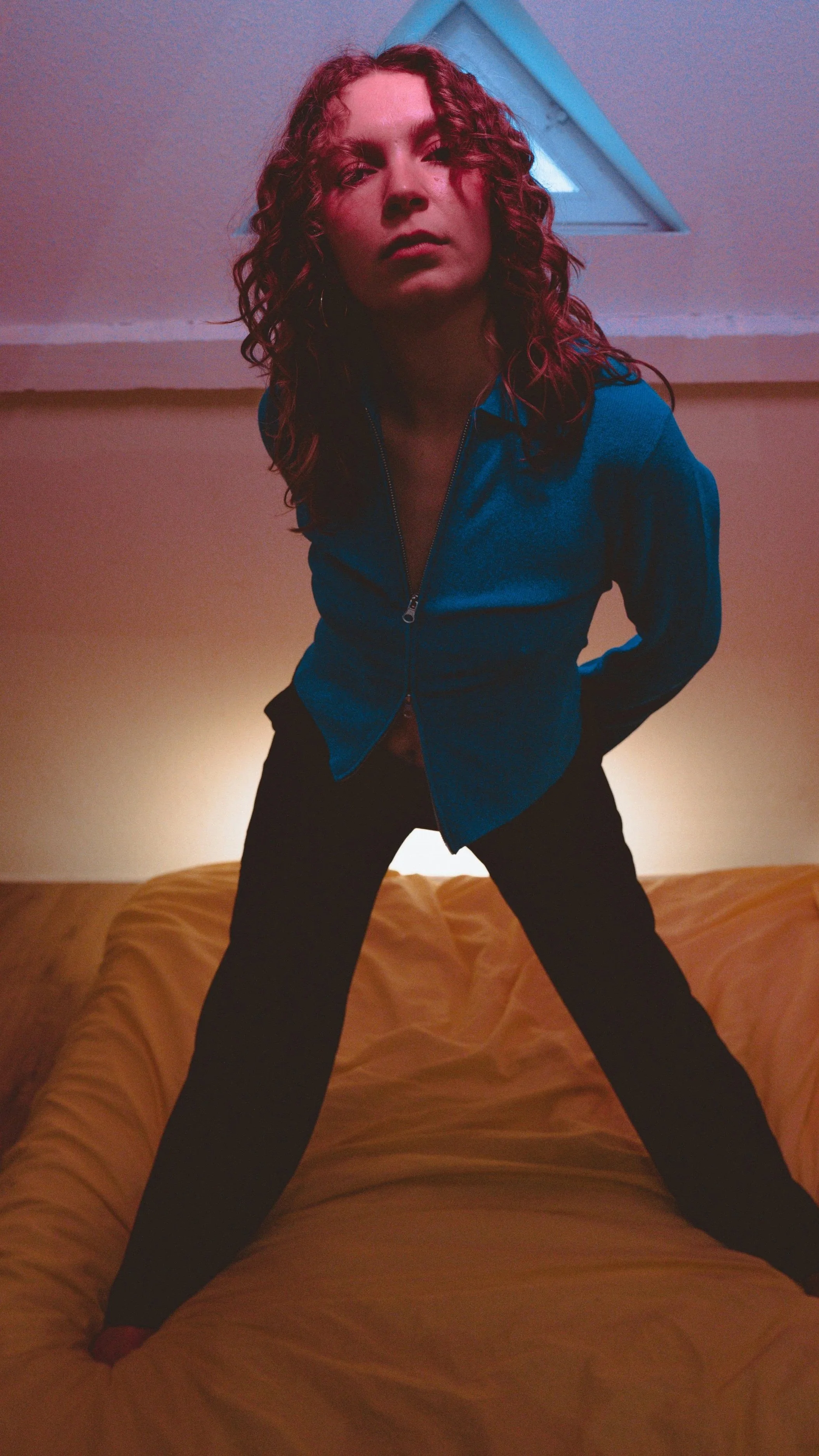 A woman with curly red hair wearing a blue zip-up jacket and black pants leans forward with legs apart on a yellow bed, looking at the camera with a serious expression.