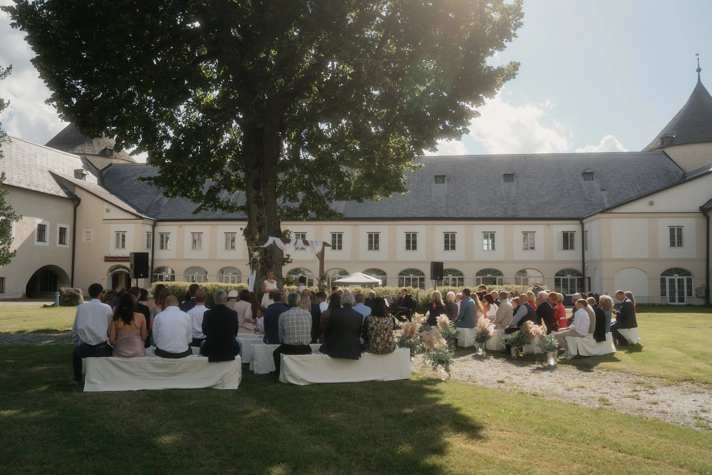 Outdoor wedding ceremony with seated guests at a historic estate or castle, under a large tree on a sunny day.