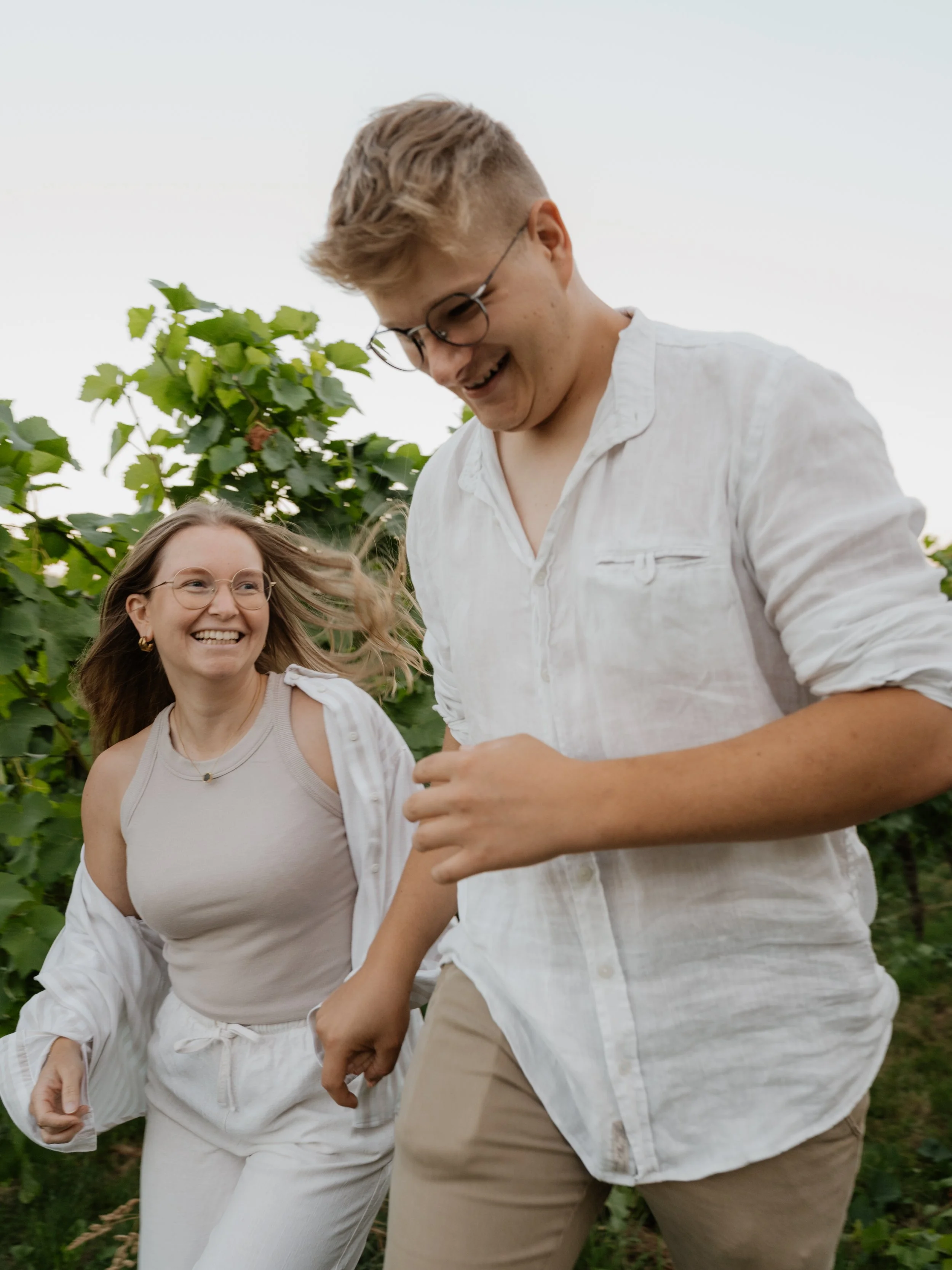 Two young people, a man and a woman, are smiling and running outdoors in a green vineyard or garden setting.