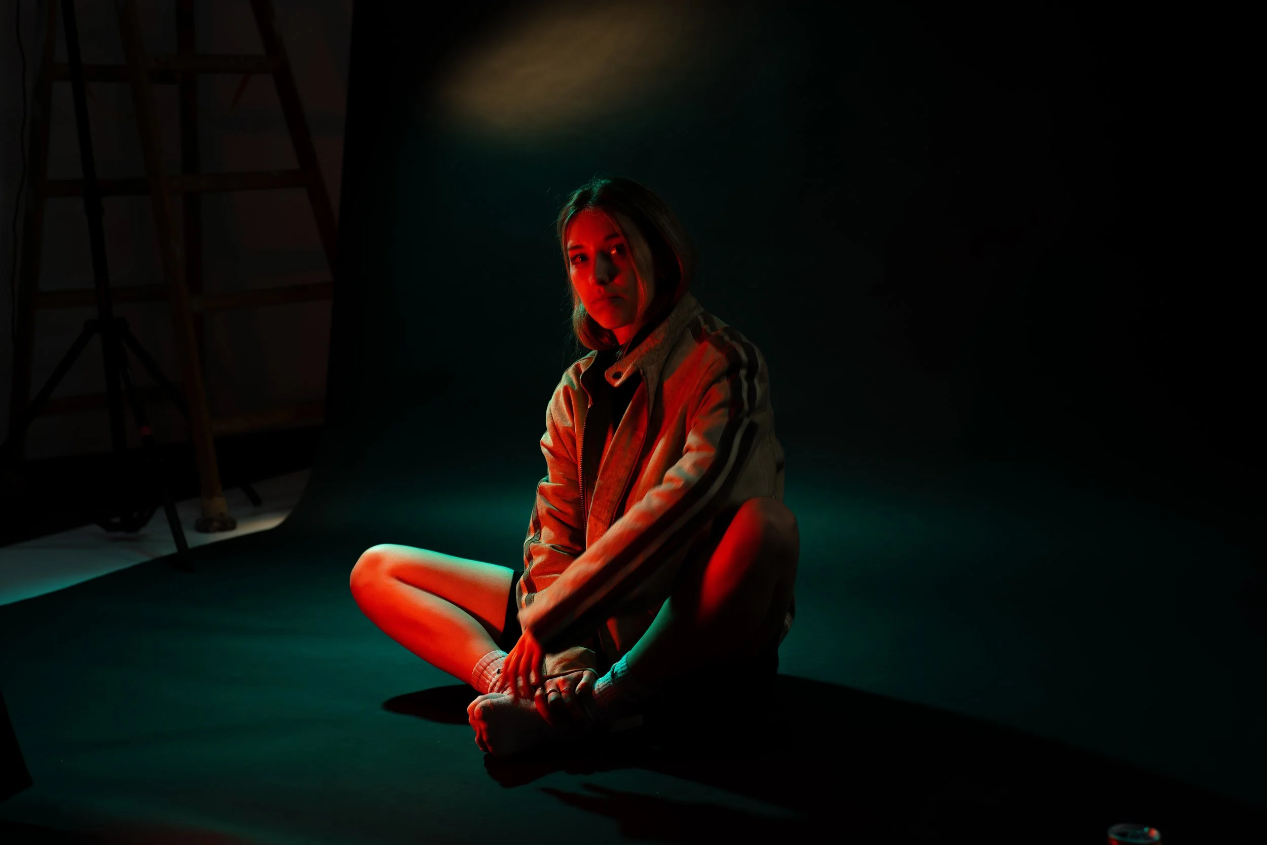 A young woman sits on the floor in a dimly lit studio, wearing a jacket and socks, illuminated by red and green lighting.