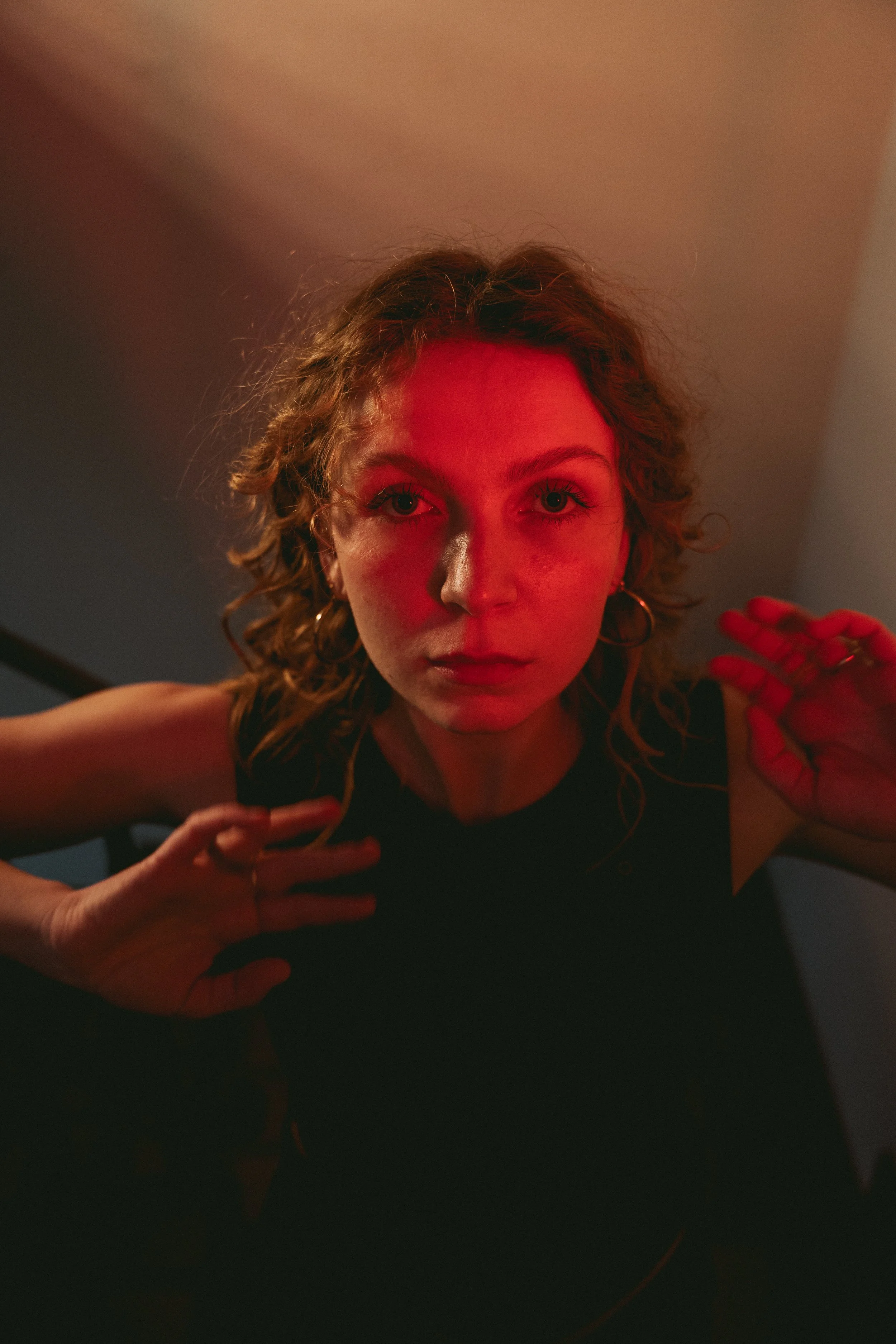 Close-up of a young woman with curly hair, wearing a black sleeveless top, under dim lighting with a red hue, looking directly into the camera with her hands near her face.