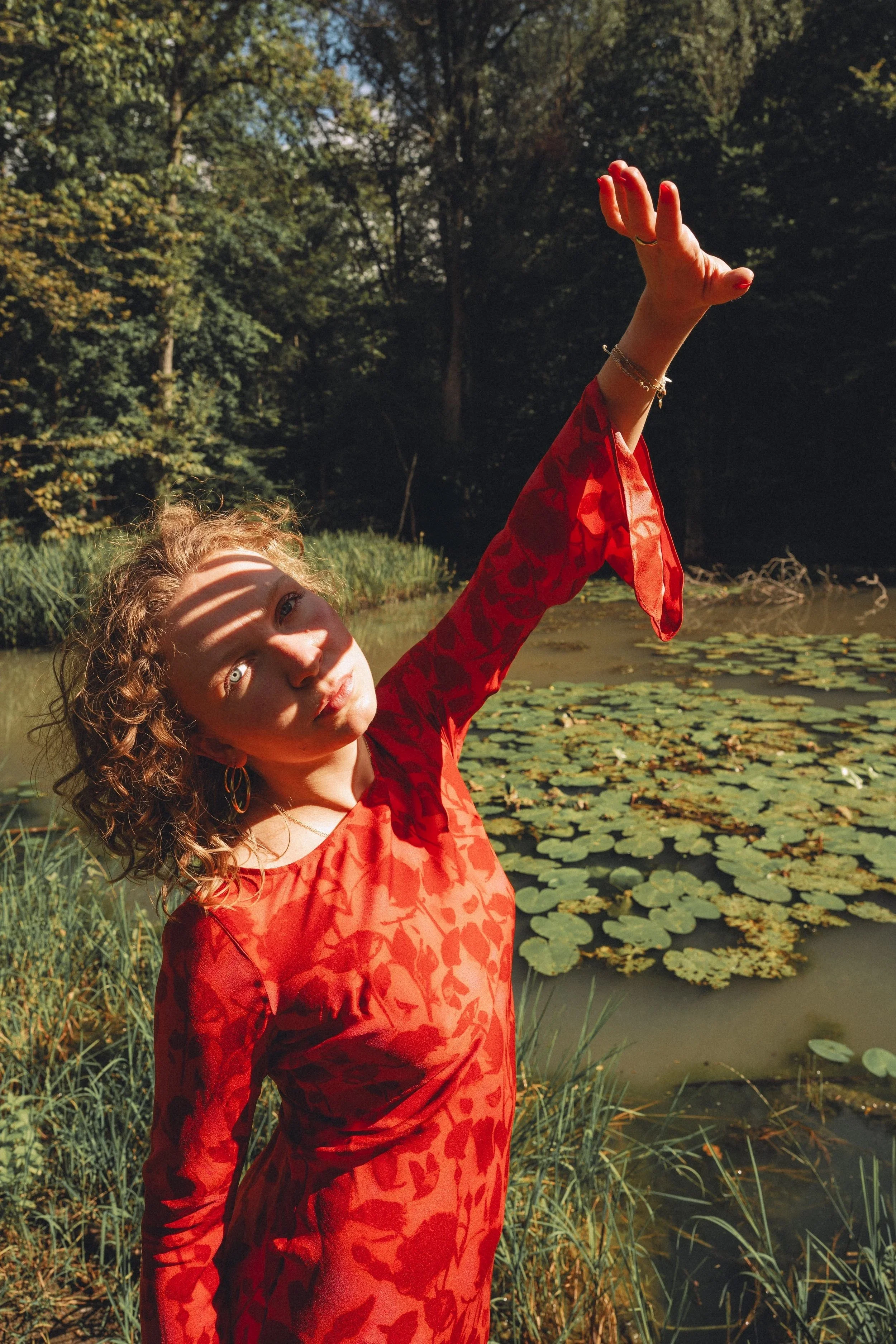 A woman with curly hair wearing a red patterned dress, standing near a pond with lily pads, reaching upwards with her left arm, in a sunlit outdoor setting surrounded by trees.
