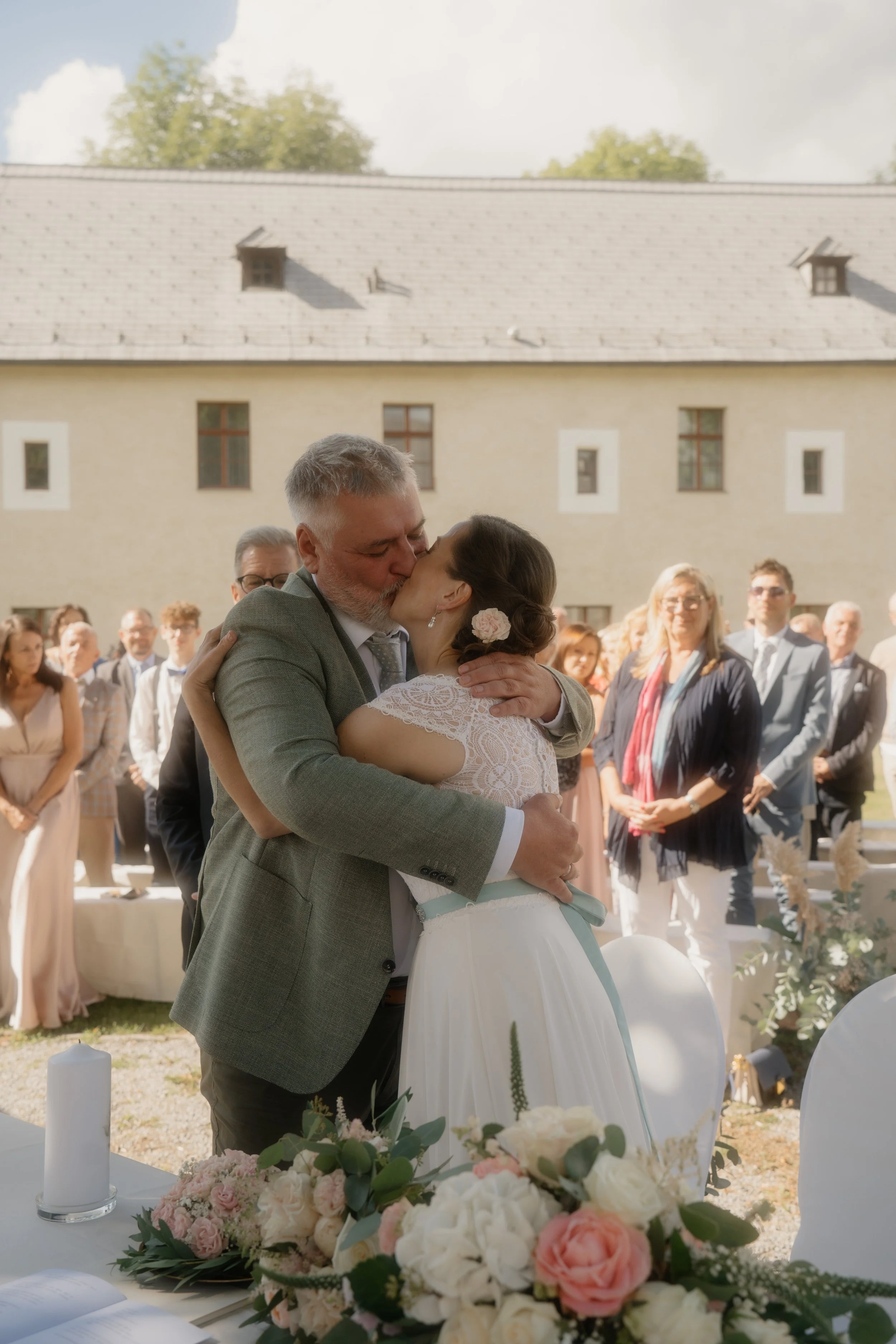 A bride and a man, possibly her father, sharing a kiss at an outdoor wedding ceremony, with guests watching in the background and floral decorations on a table in the foreground.