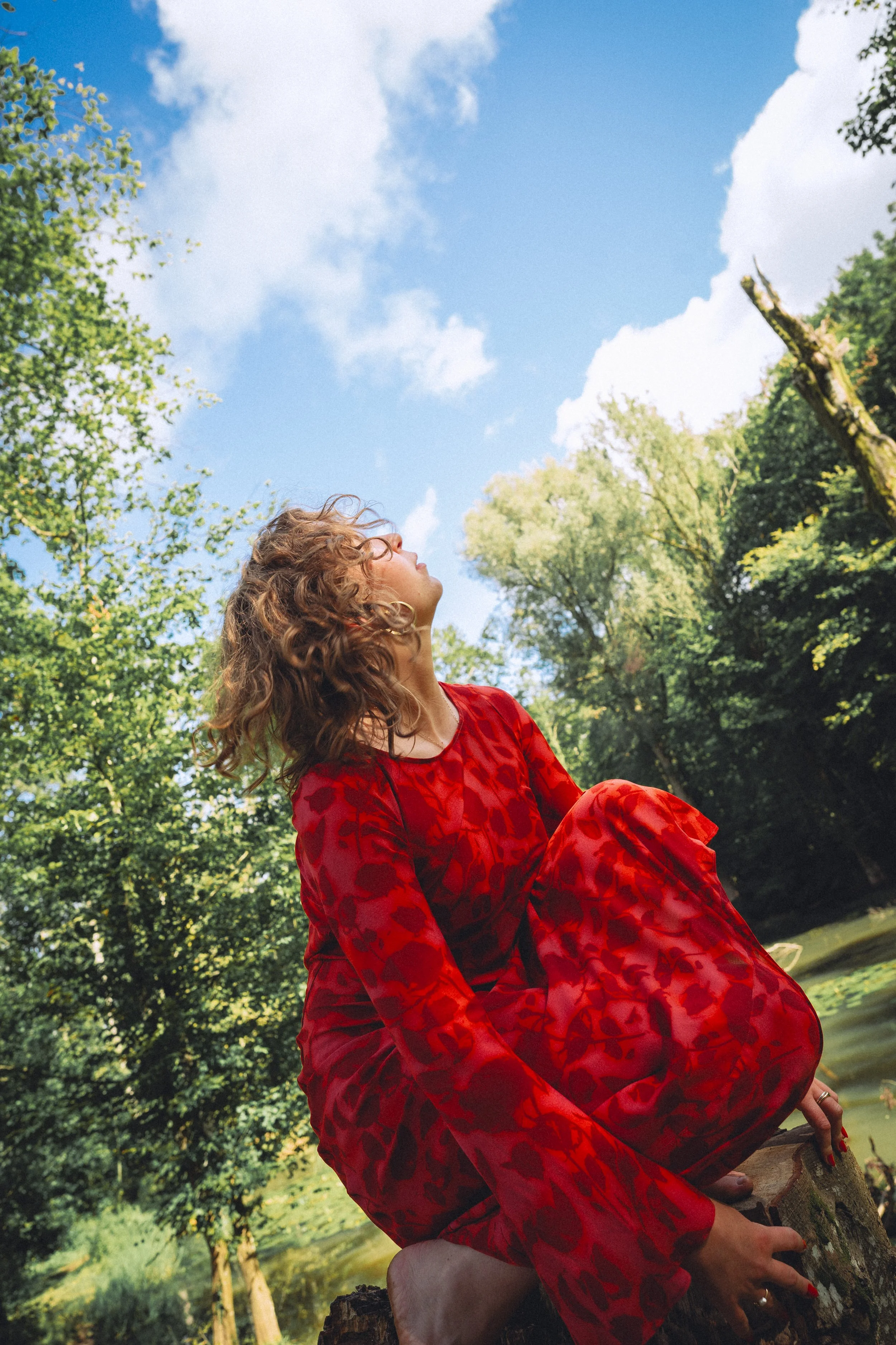 A woman with curly hair in a red dress sitting on a tree stump by a river, looking up at the sky, with trees and a blue sky with clouds in the background.
