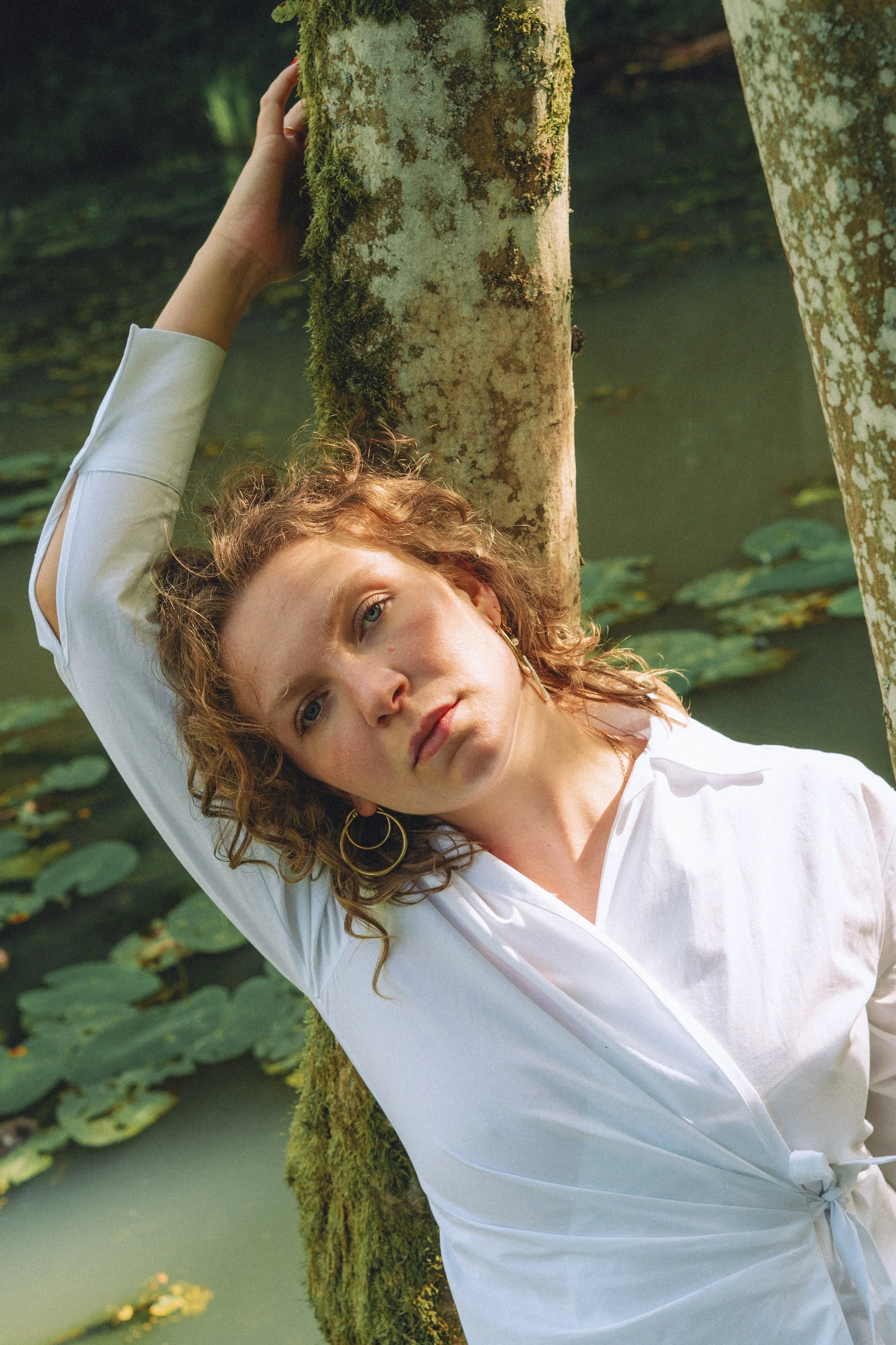 A woman with curly red hair and gold hoop earrings leaning against a moss-covered tree near a lily pad pond, wearing a white shirt.