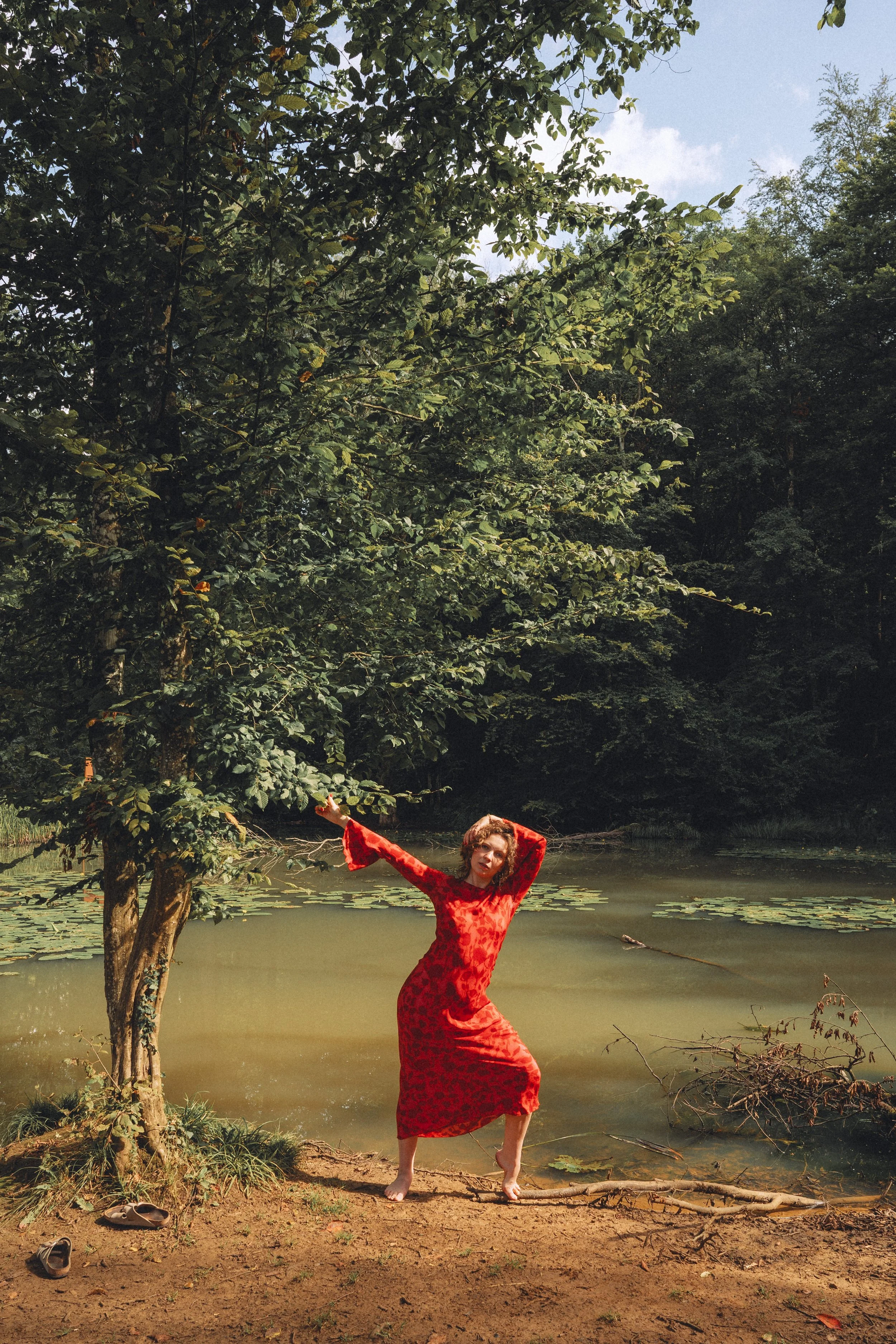 A woman in a red dress standing barefoot by a pond, surrounded by trees with green foliage.