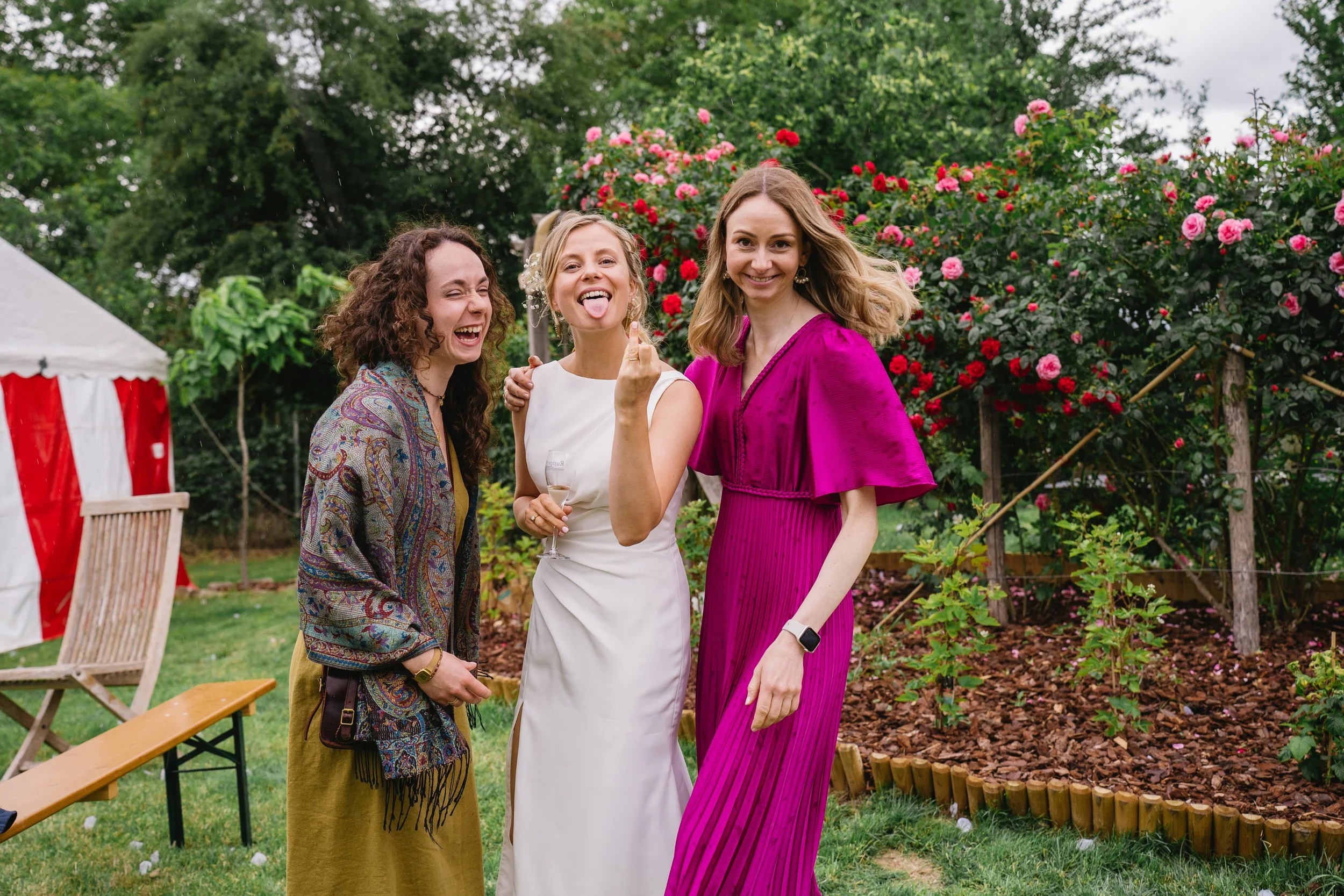 Three women smiling and posing outdoors during a garden event, with roses and trees in the background, one holding a glass of champagne.