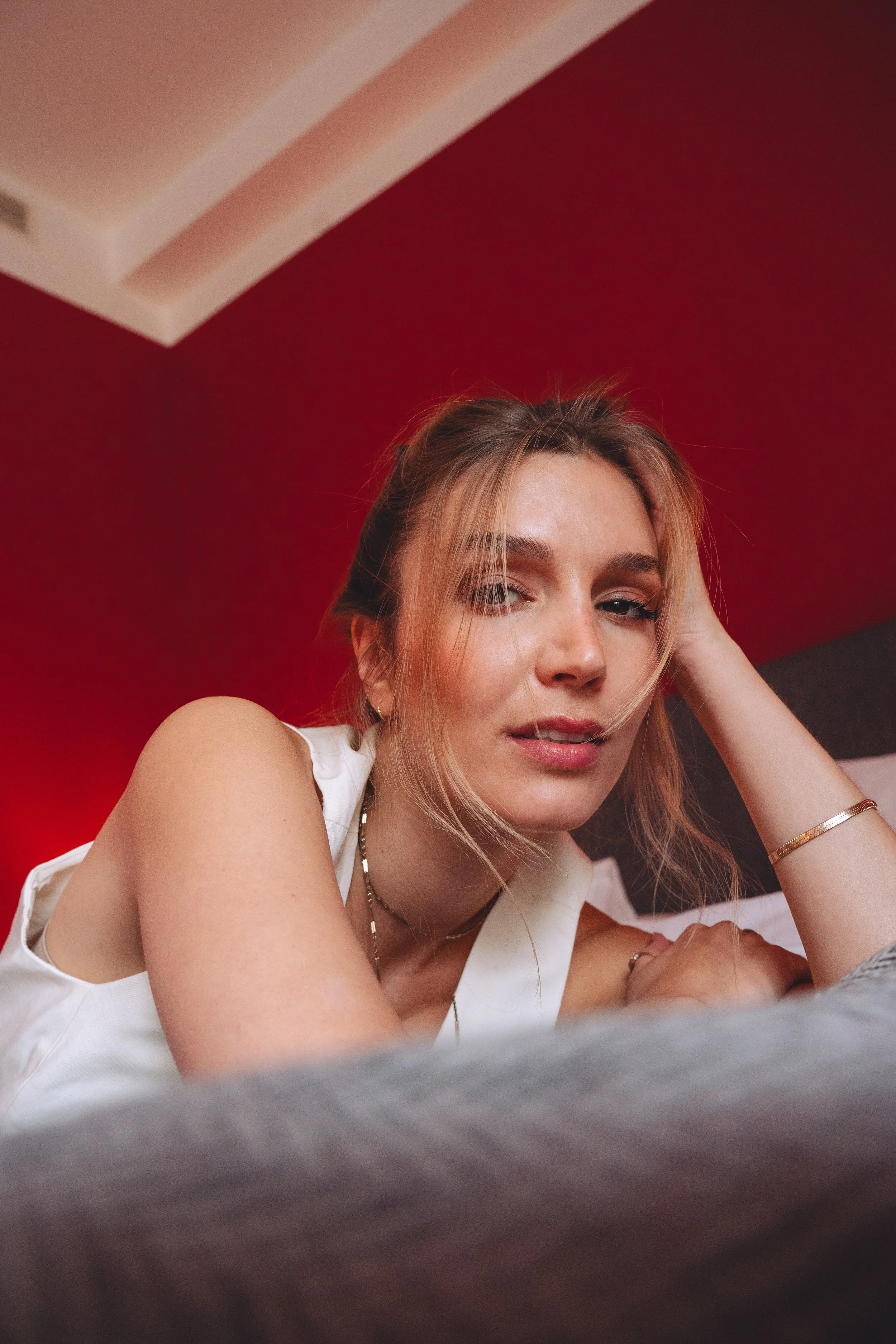 Young woman lying on a bed, resting her head on her hand, with a red wall behind her and a white ceiling above.