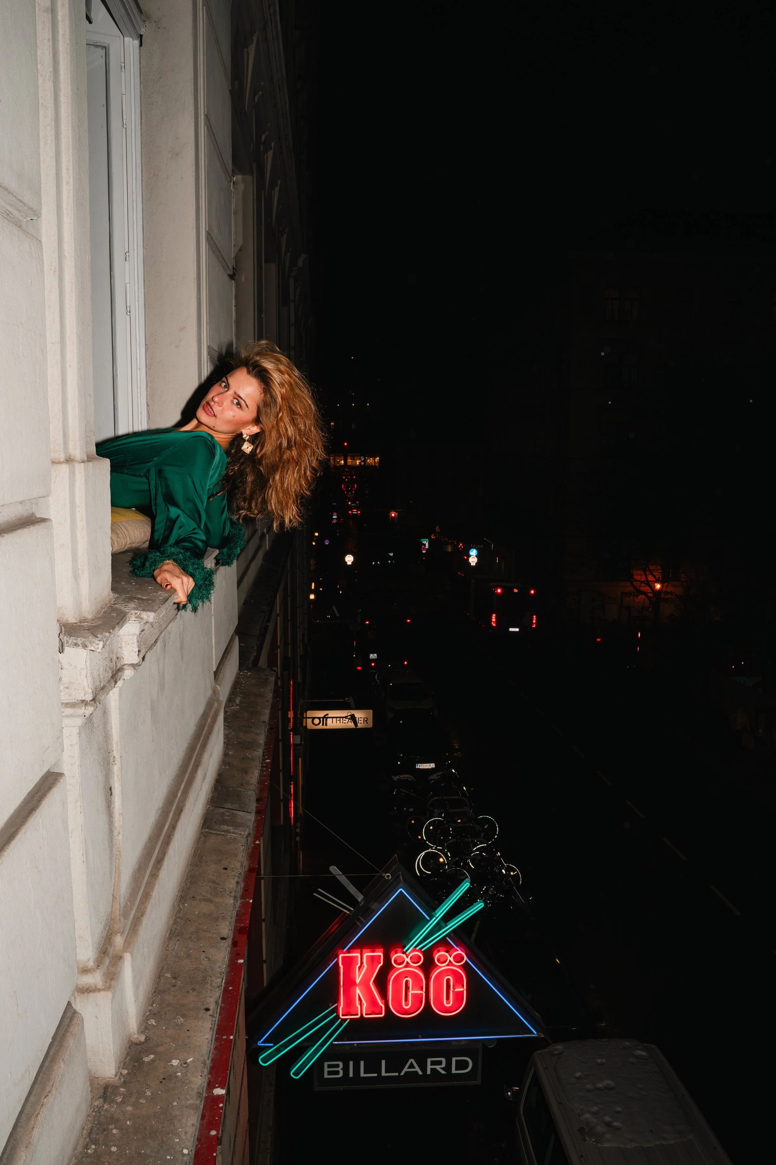 A woman with curly red hair in a green satin blouse leaning out of a window at night, with a city street below and neon signs including one that reads 'KÖÖ' and 'Billard'.