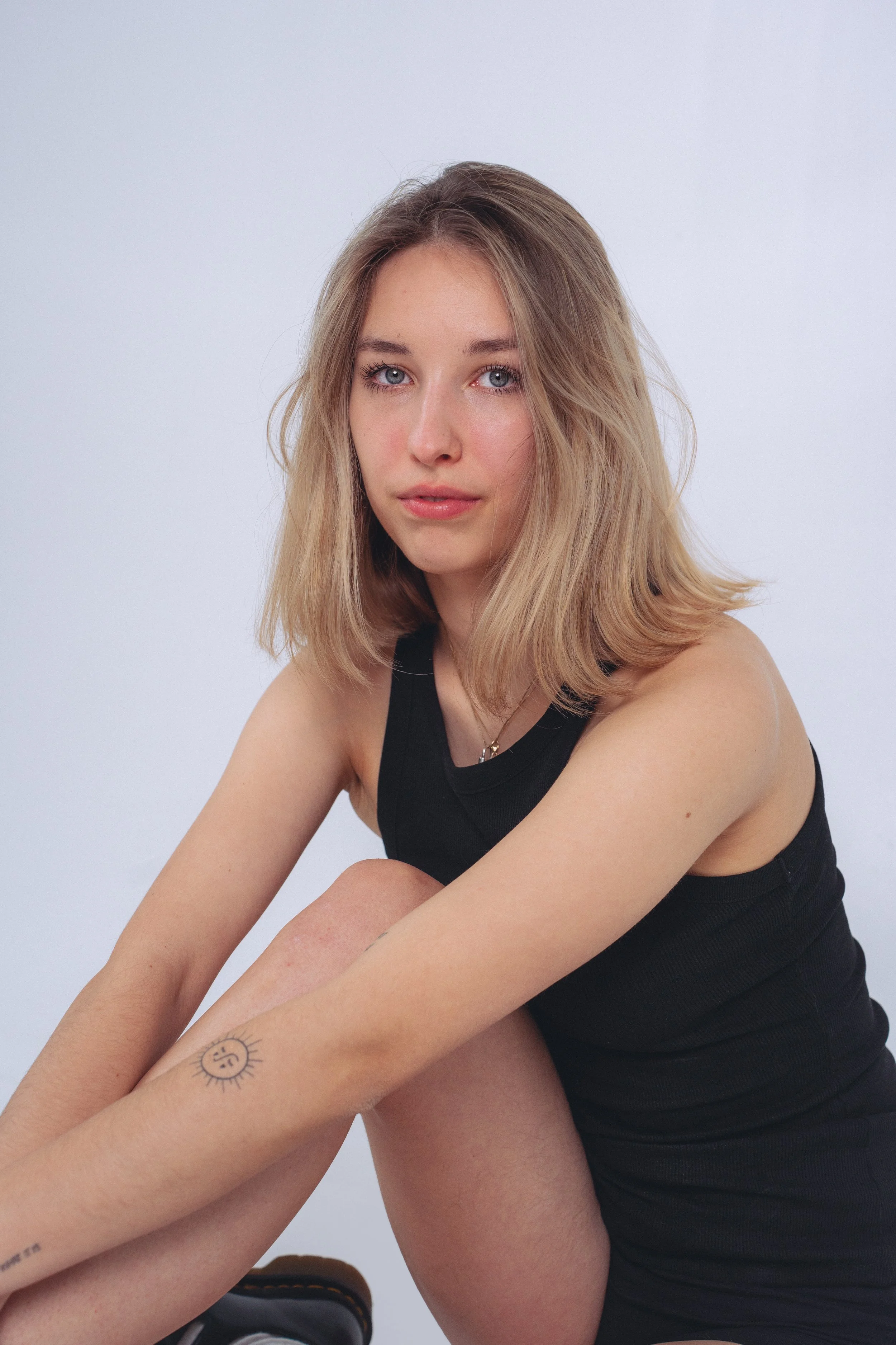 A young woman with shoulder-length blond hair and blue eyes, wearing a black sleeveless top, sitting against a plain light background.
