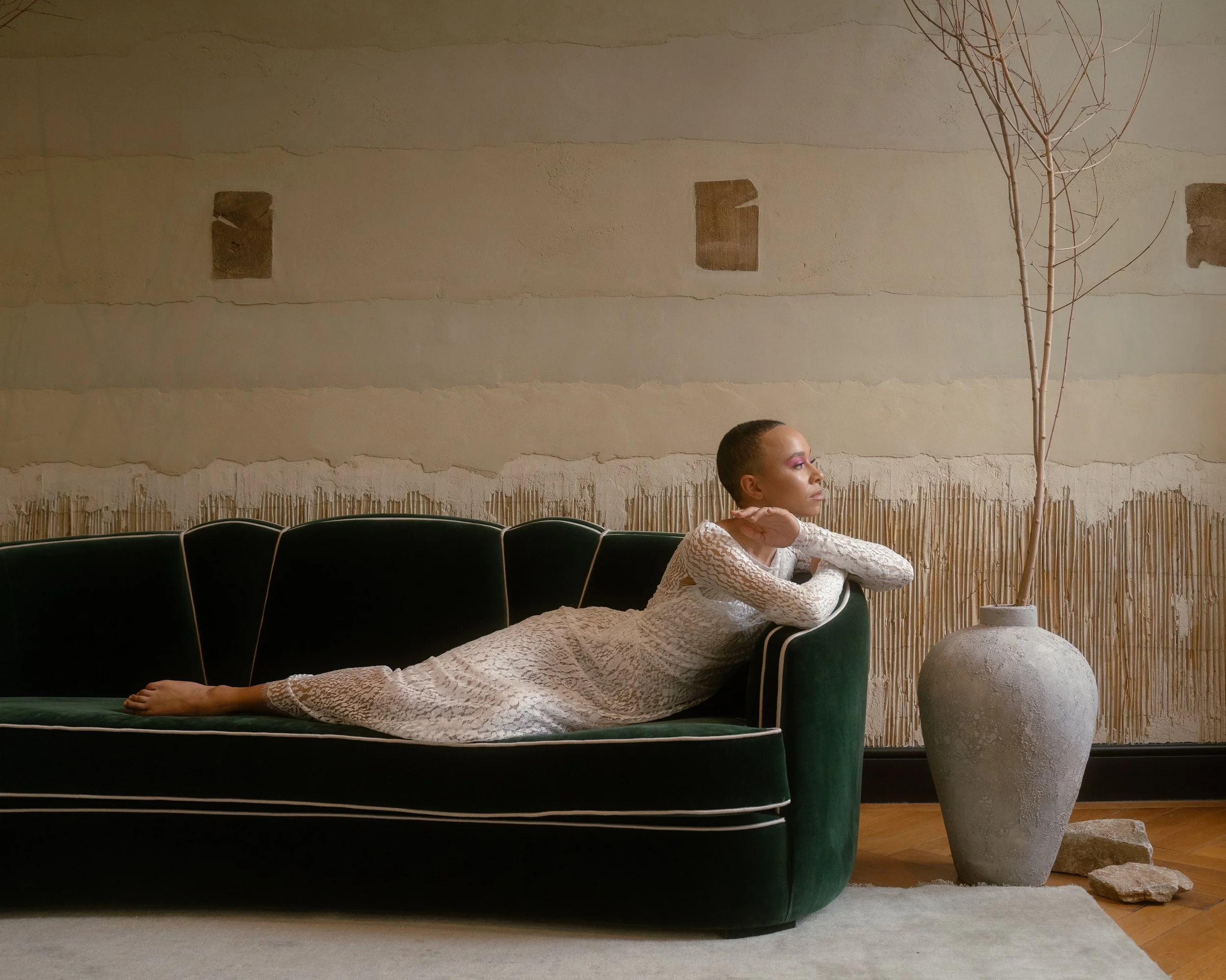 A woman in a white lace dress lying on a green velvet sofa, resting her chin on her crossed arms, with a large potted tree nearby, in a minimalist room with a textured beige wall.