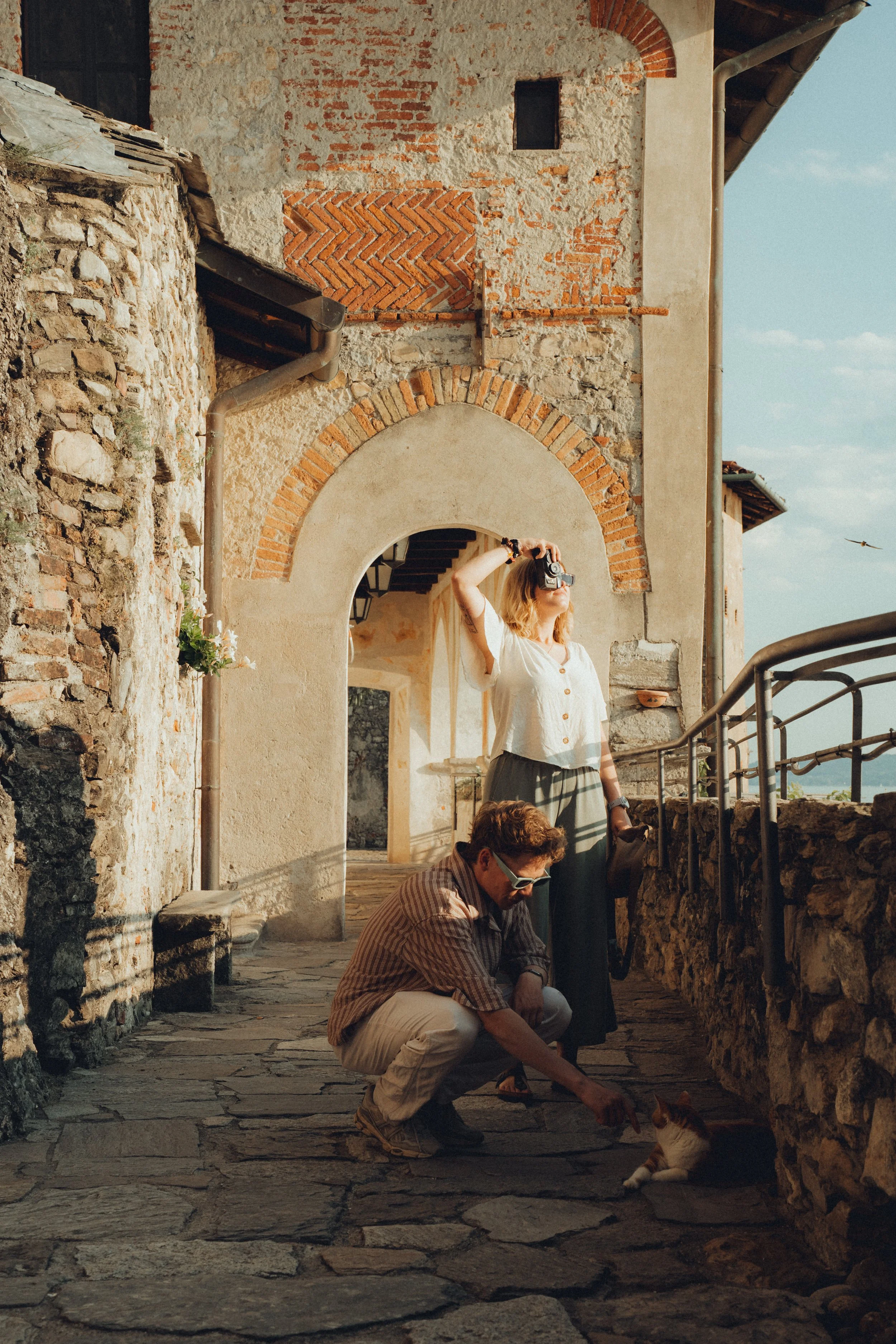 Two people, a woman standing and a man crouching, near a stone wall with an arched doorway, with a cat lying on the ground, all in a rustic outdoor setting.