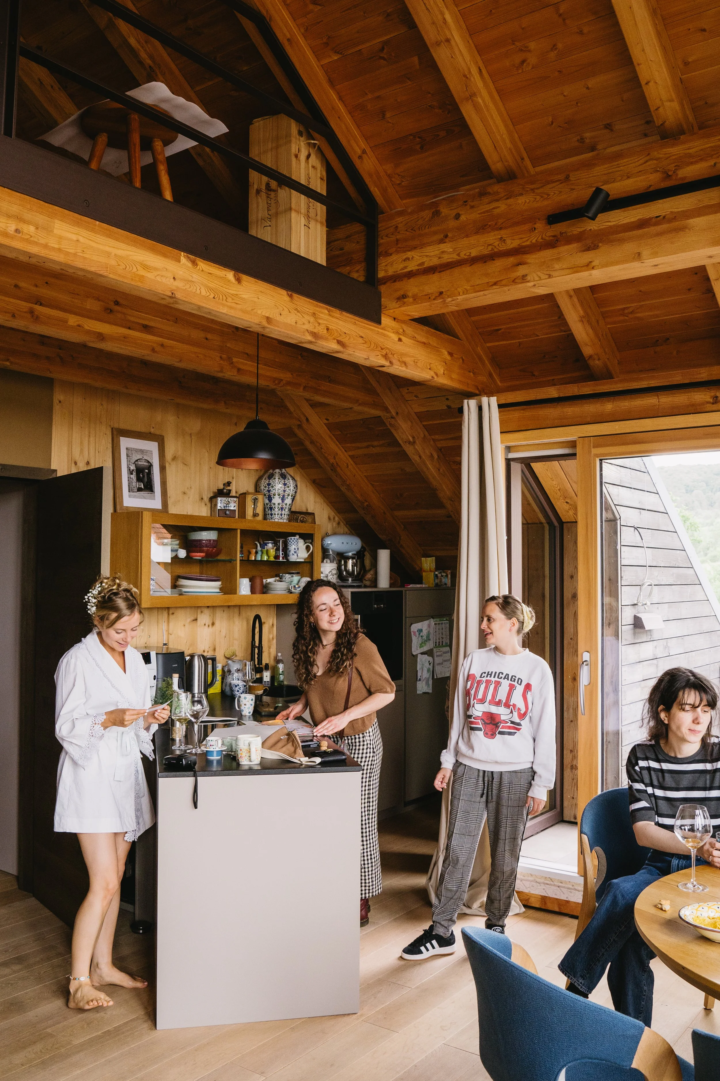 Four women in a cozy, wood-paneled kitchen and dining area socializing and preparing food, with a lofted space above.