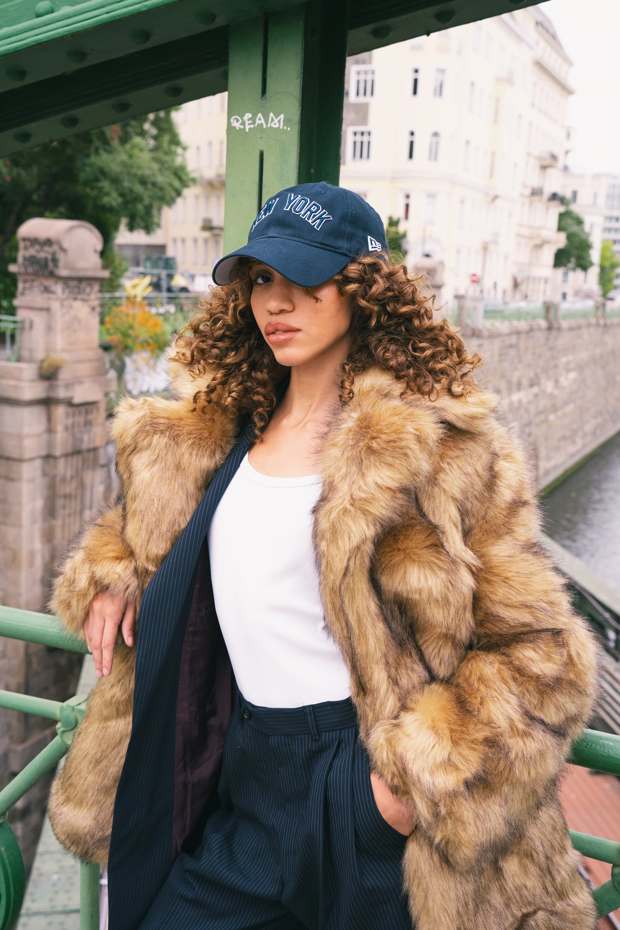 A woman with curly hair wearing a New York Yankees cap, a fur coat, a white shirt, and pinstriped pants, standing on a bridge over a canal in an urban area with buildings and greenery in the background.