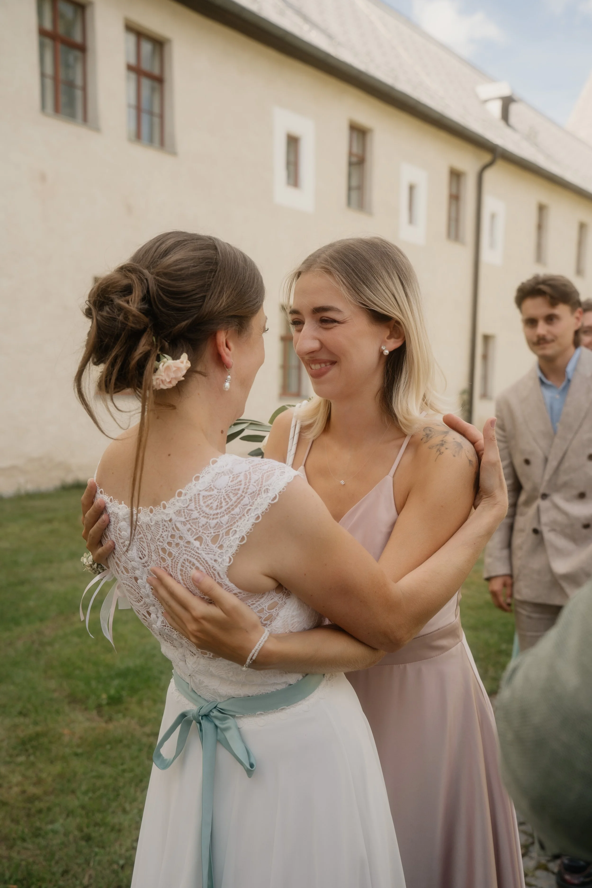 Two women embracing at a wedding, one in a white dress with lace detail and a light blue sash, and the other in a blush-colored dress, smiling at each other. A man in a beige suit stands in the background, outside a light-colored building.