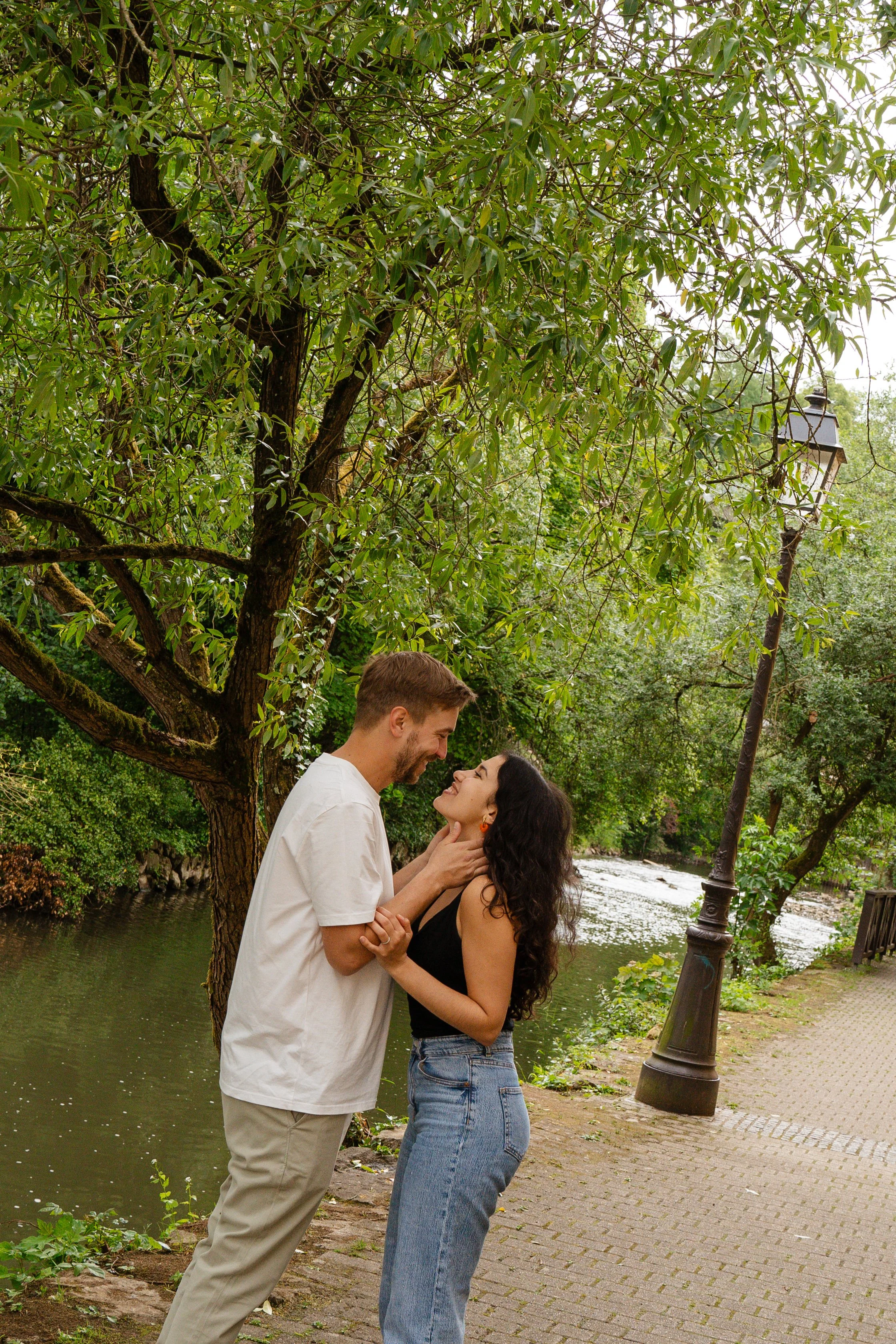 A couple standing on a paved walkway next to a lake or river, smiling and looking at each other, surrounded by greenery and trees, with a lamppost nearby.