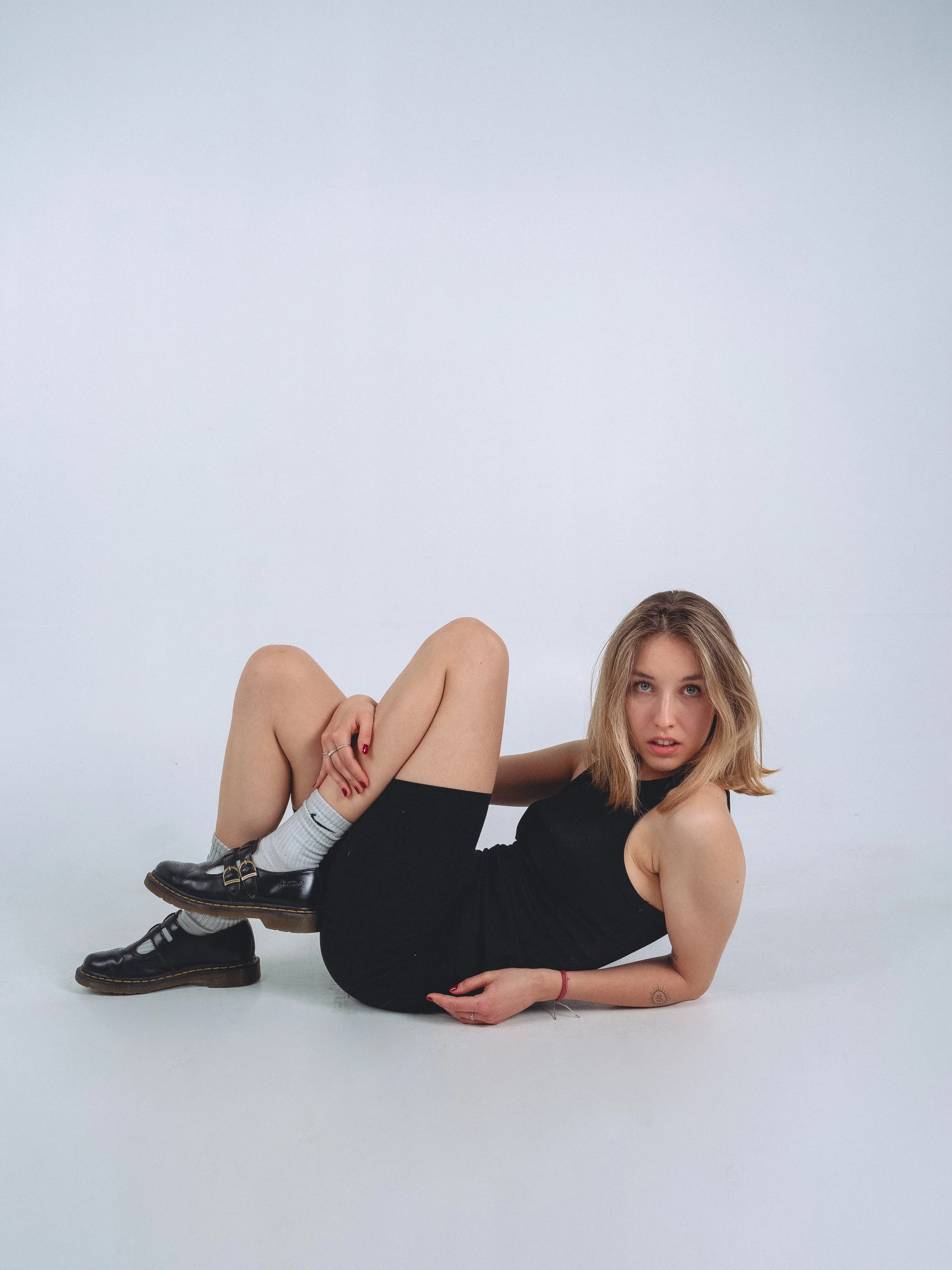 A young woman with blonde hair, wearing a black dress, grey socks, and black Mary Jane shoes, lying on a white surface against a plain white background, looking at the camera.