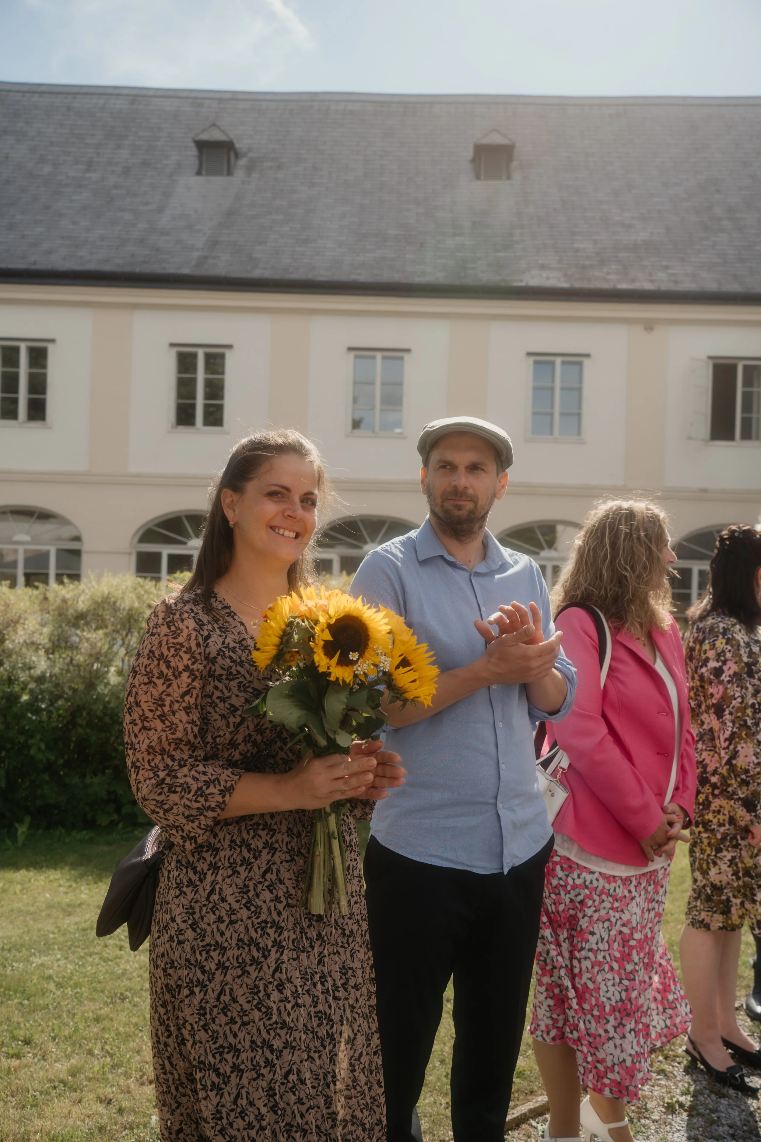 A woman holding a bouquet of sunflowers standing next to a man clapping at an outdoor gathering.