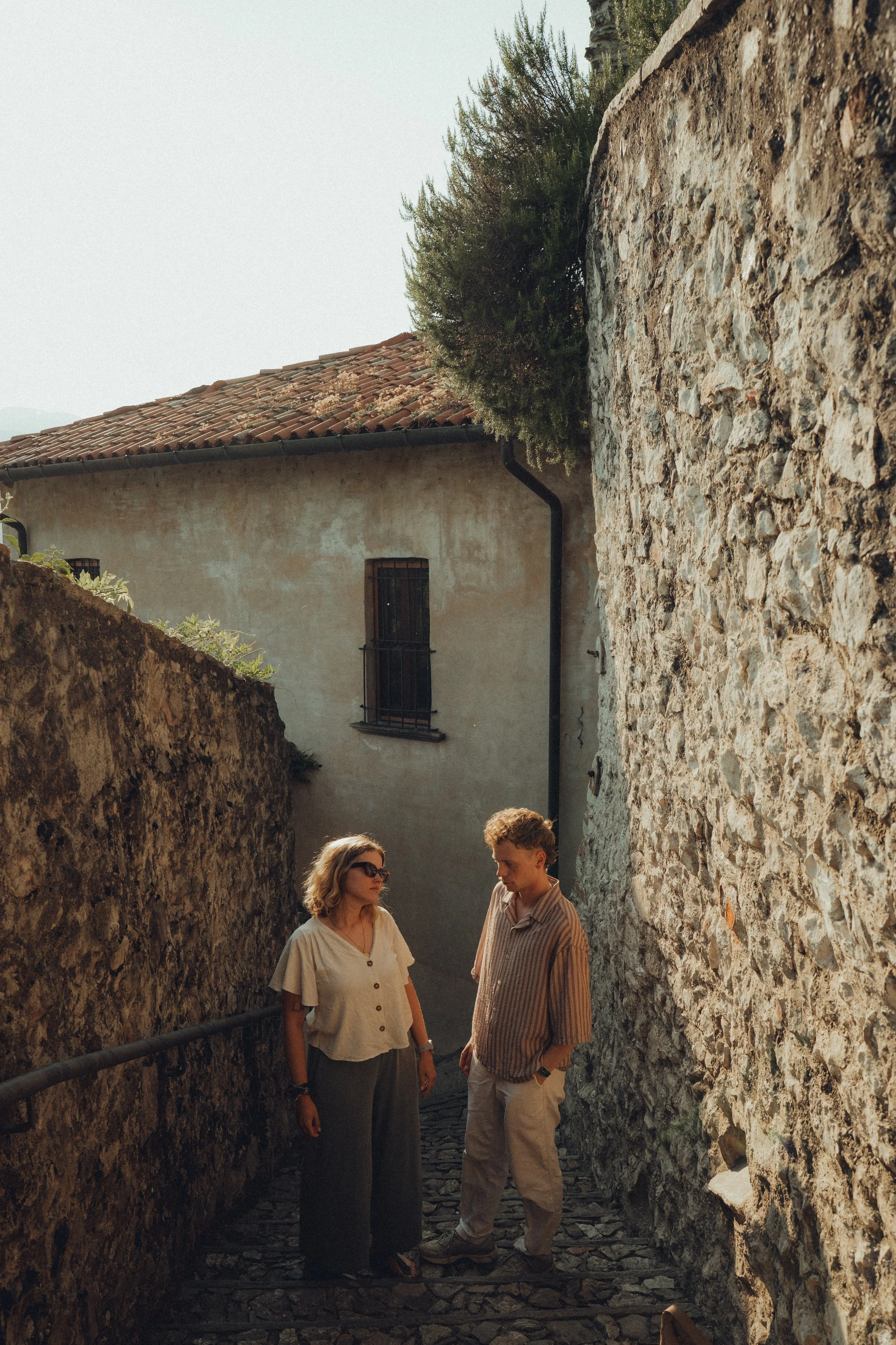 Two people standing on a narrow cobblestone alleyway between stone walls, engaged in conversation on a sunny day.