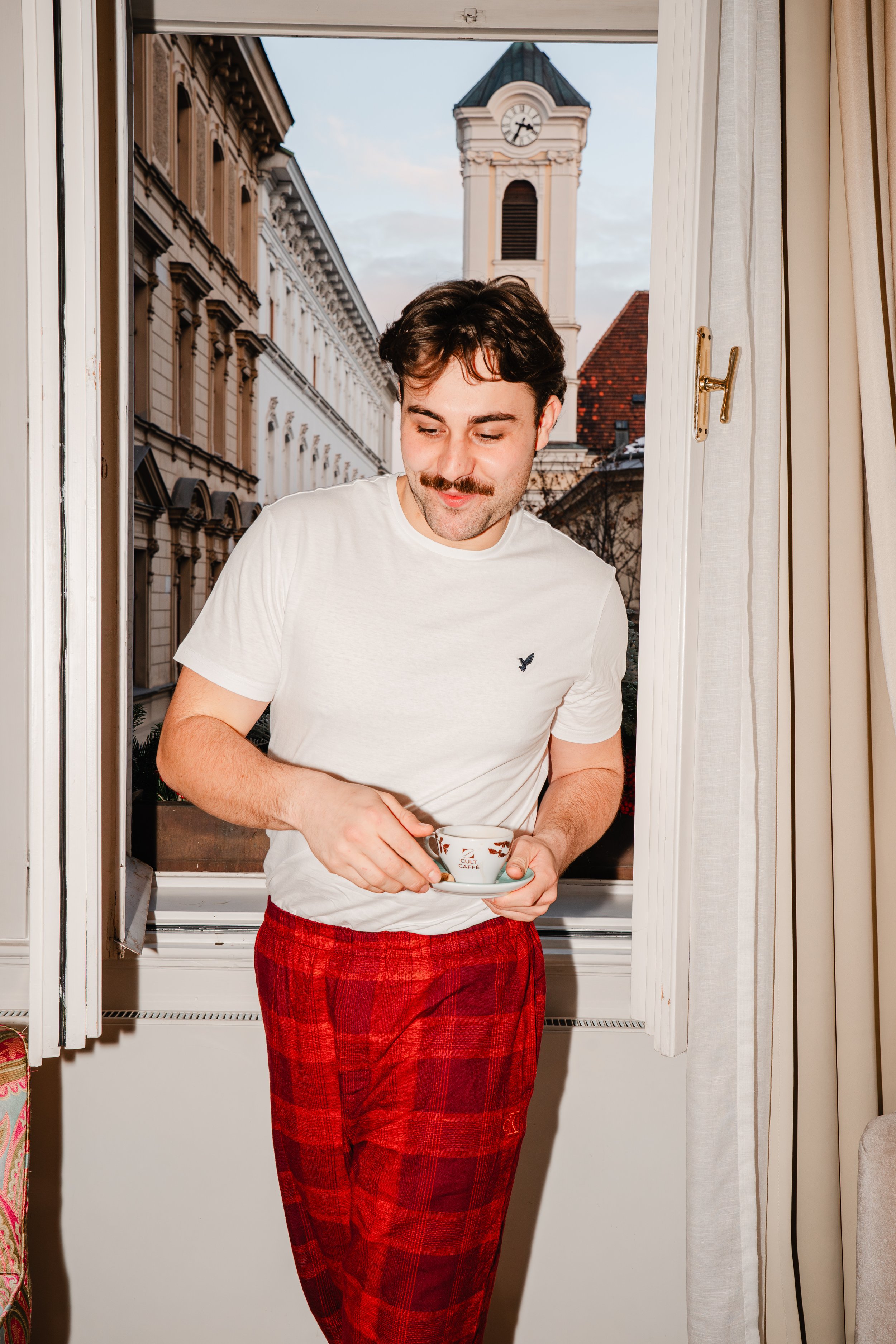 A young man with a mustache and dark hair, wearing a white T-shirt and red plaid pajama pants, holding a small teacup and looking down, standing in front of an open window with a cityscape and clock tower in the background.