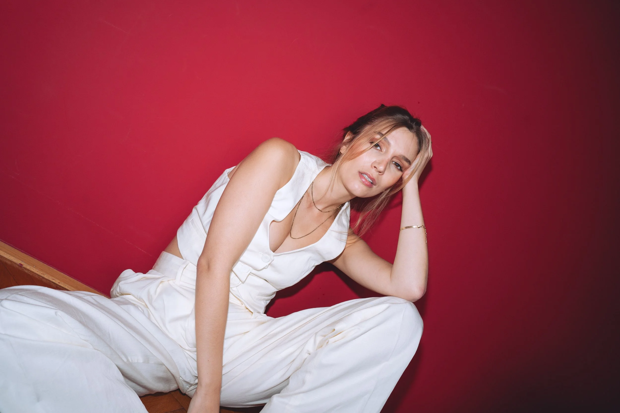 A young woman with light brown hair and light skin, wearing a white sleeveless top and white pants, sitting on the floor against a red wall. She is resting her head on her hand, with a relaxed and slightly sultry expression.