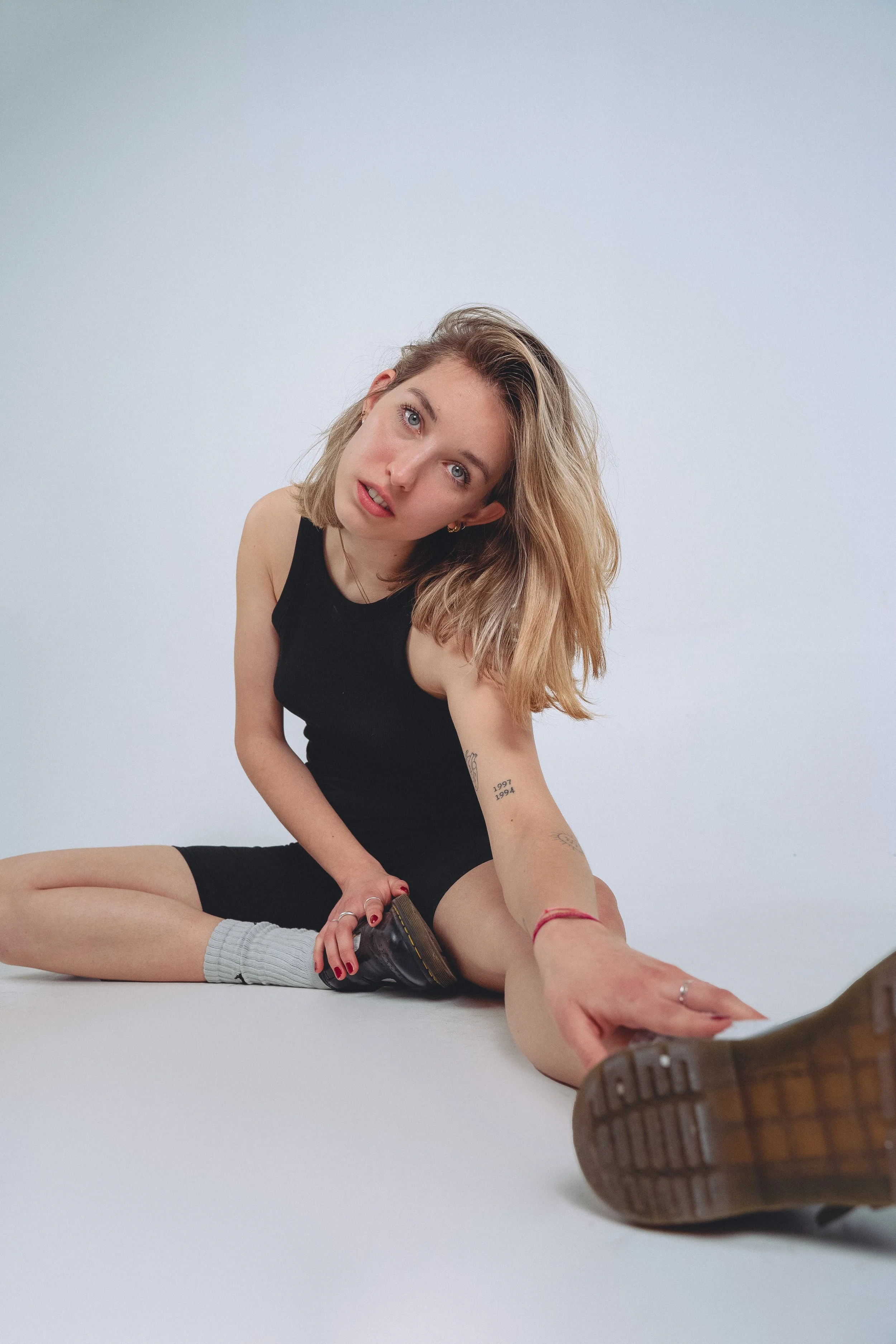 A young woman with blonde hair, wearing a black tank top, black shorts, gray socks, and brown shoes, sits on the floor stretching her leg forward while looking at the camera with a relaxed expression.