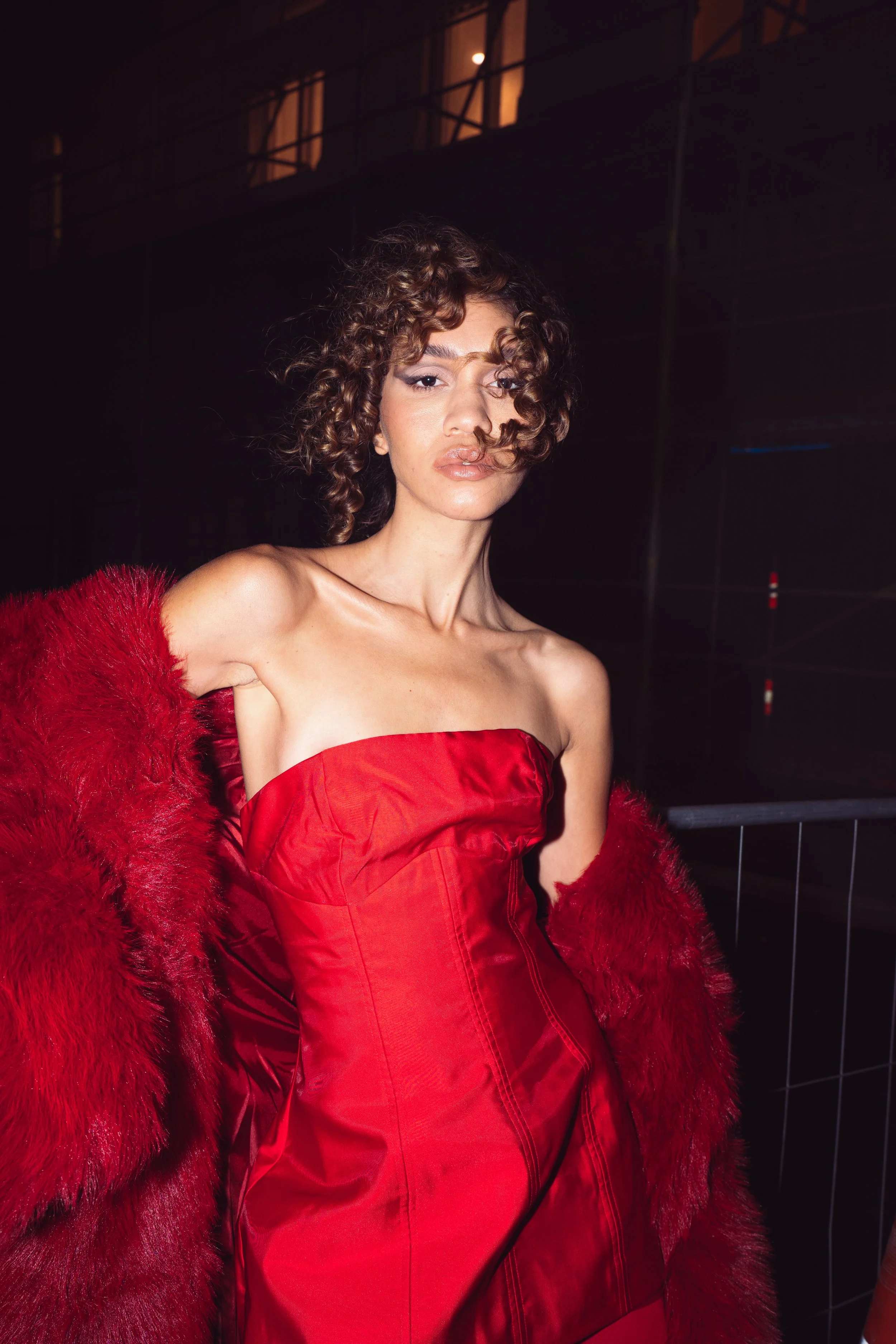 A woman with curly hair wearing a strapless red dress and a red fur coat at night.