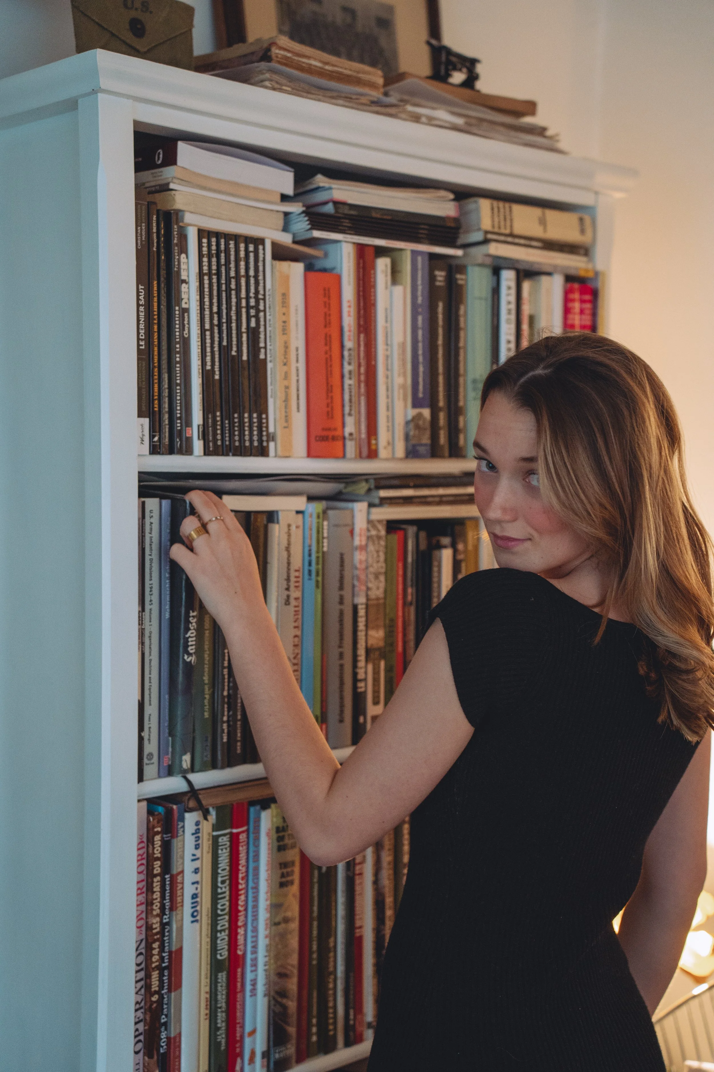 A woman with light brown hair wearing a black dress is standing next to a white bookshelf filled with books, reaching to pick a book from the middle shelf.