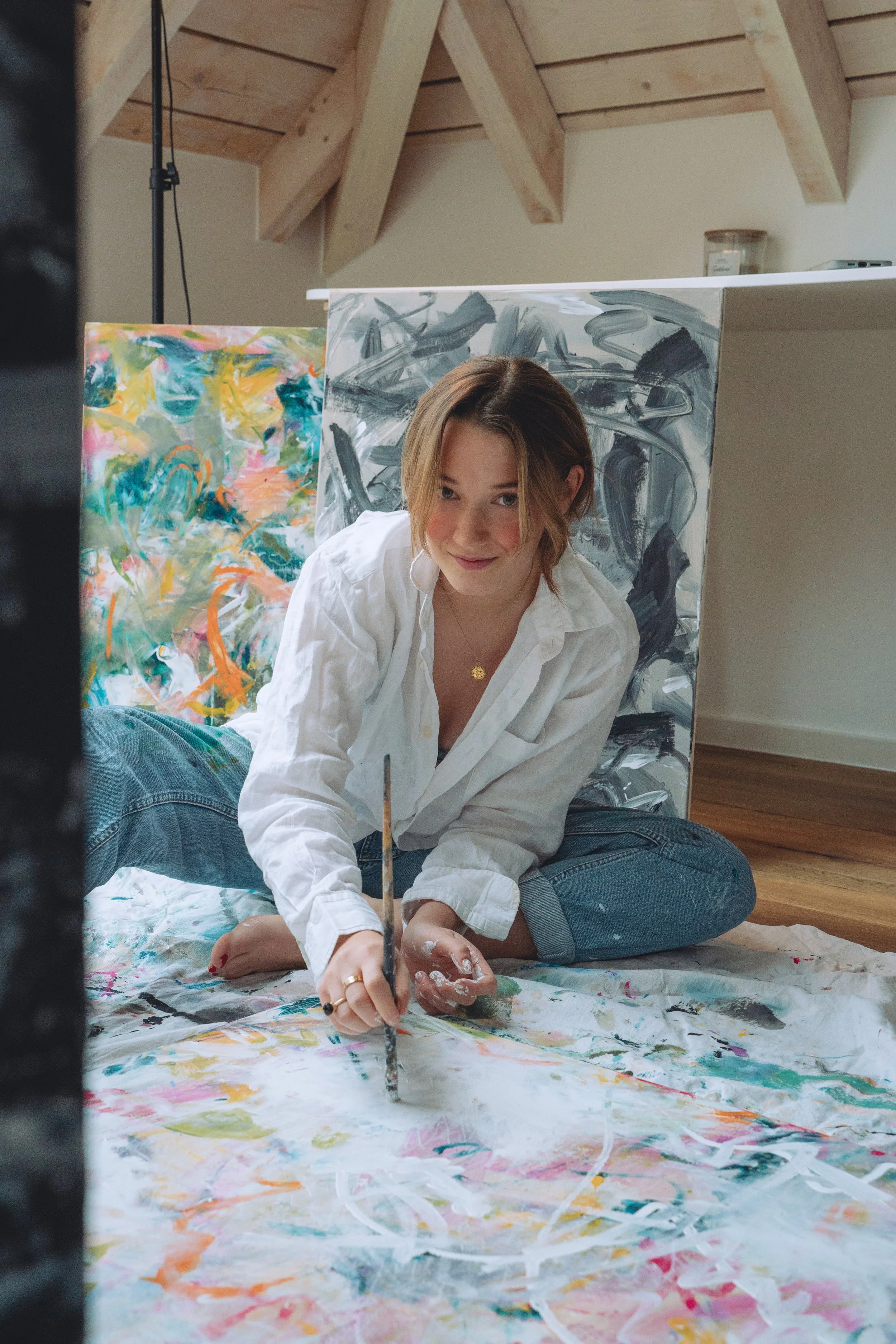 A young woman sitting cross-legged on the floor, painting abstract artwork on a large canvas. She has short brown hair, is wearing a white shirt and jeans, and is smiling at the camera.
