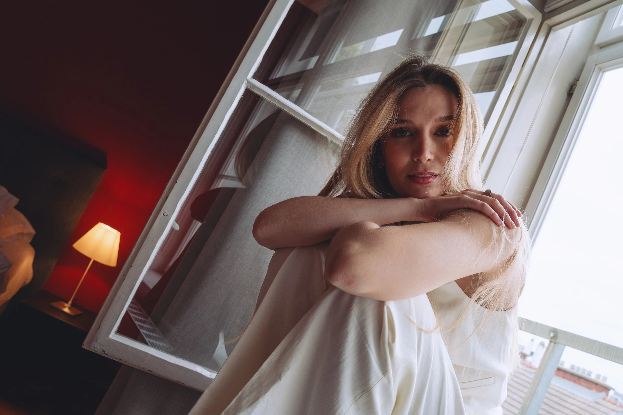 A young woman with blonde hair sitting by a large window, looking at the camera, with sunlight illuminating her face. Behind her, a balcony railing and a partly cloudy sky are visible.