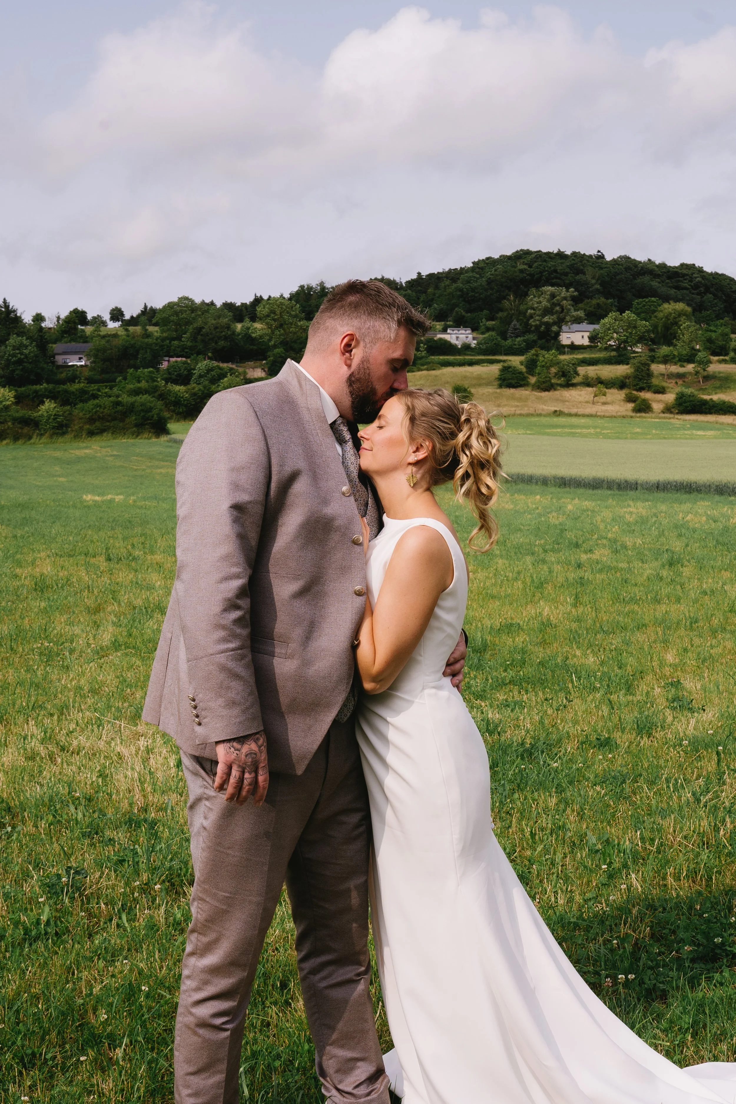 A couple dressed in wedding attire standing in a green field, with the man gently kissing the woman's forehead as they embrace, and hills and trees in the background.