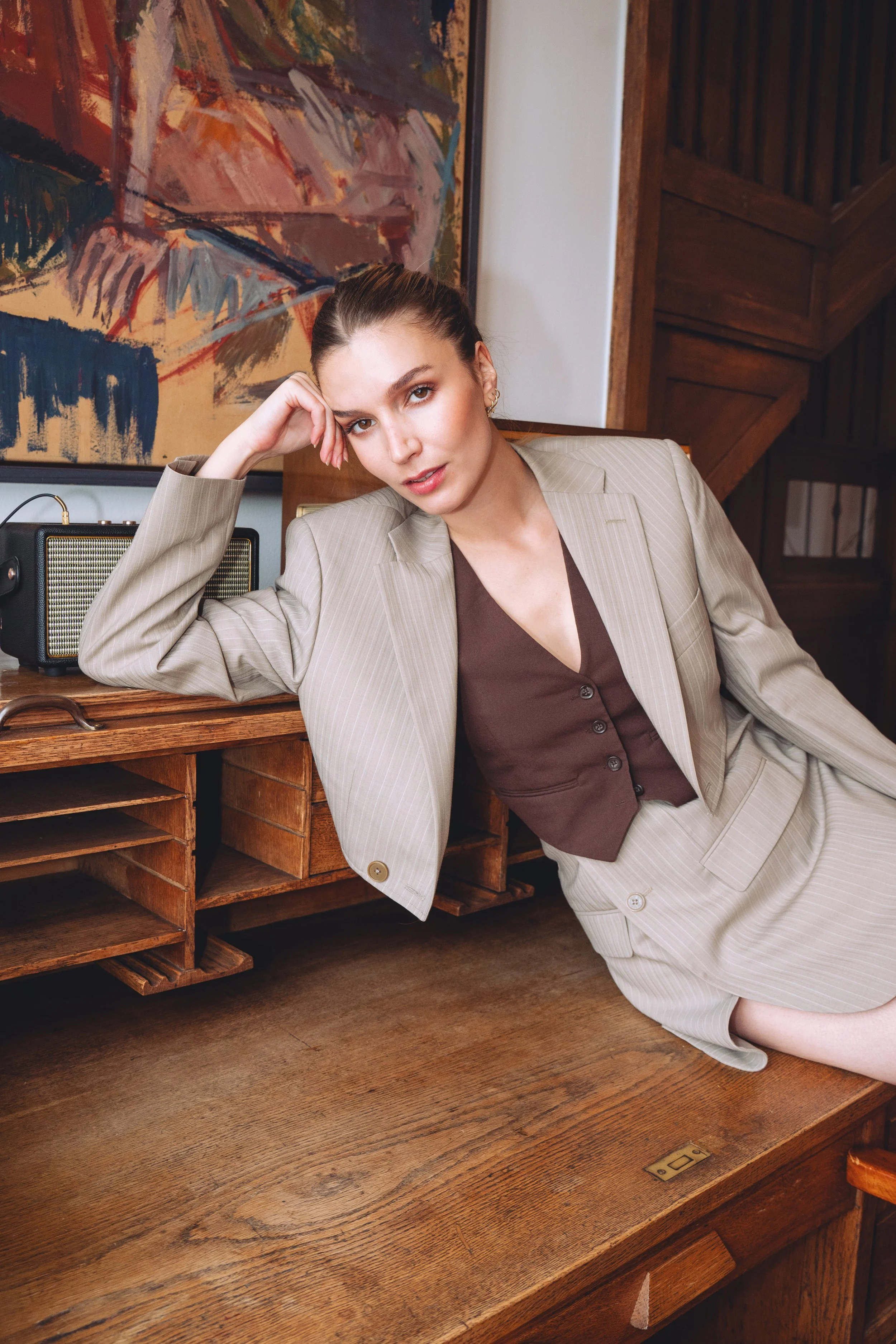 A woman in a beige pinstriped suit with a brown vest sitting on a wooden desk in a room with modern art and wooden details.