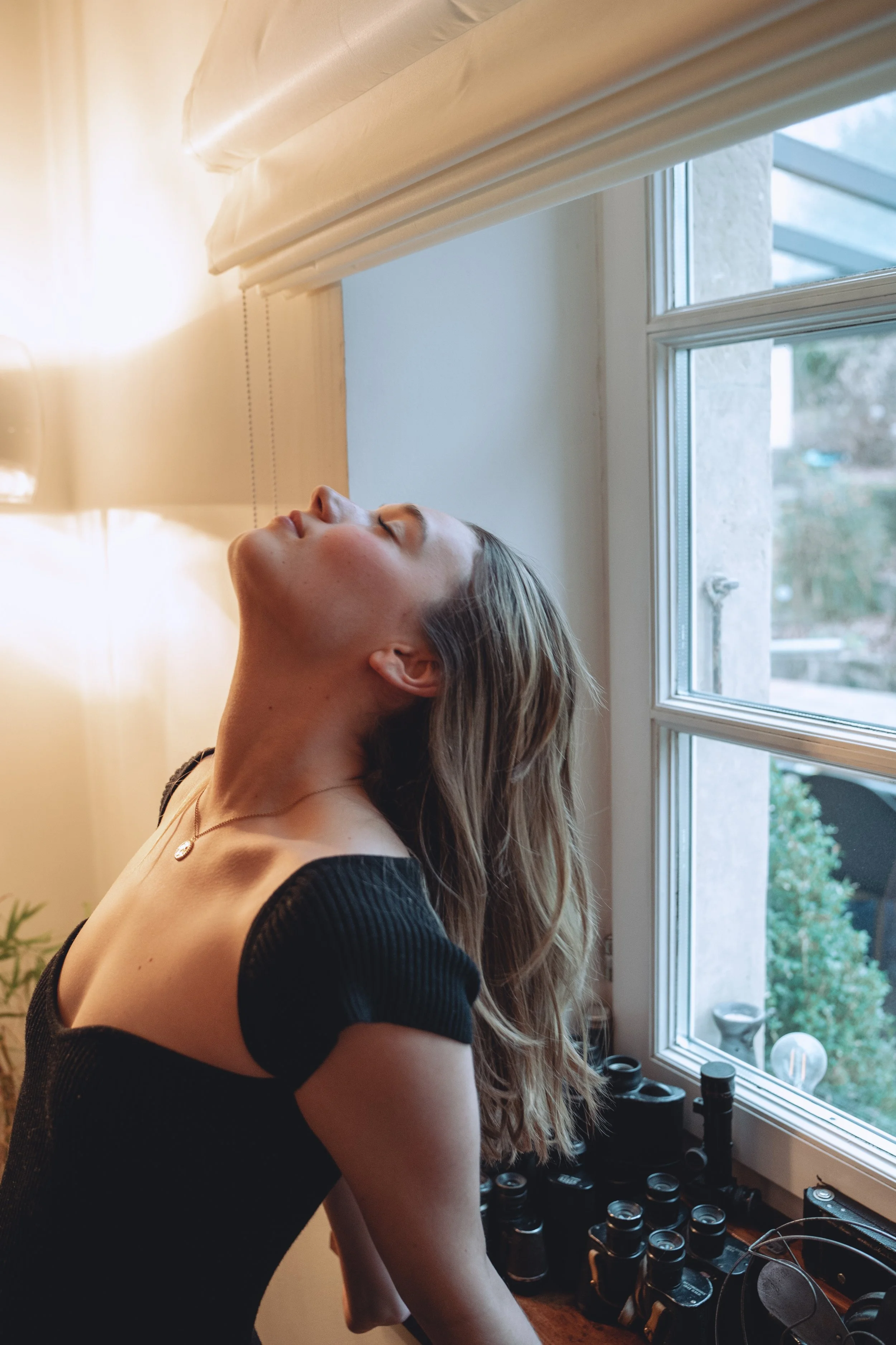 A woman with long hair, wearing a black off-shoulder top and a necklace, leaning her head back with her eyes closed near a window with beige blinds, with perfume bottles on a wooden surface nearby.