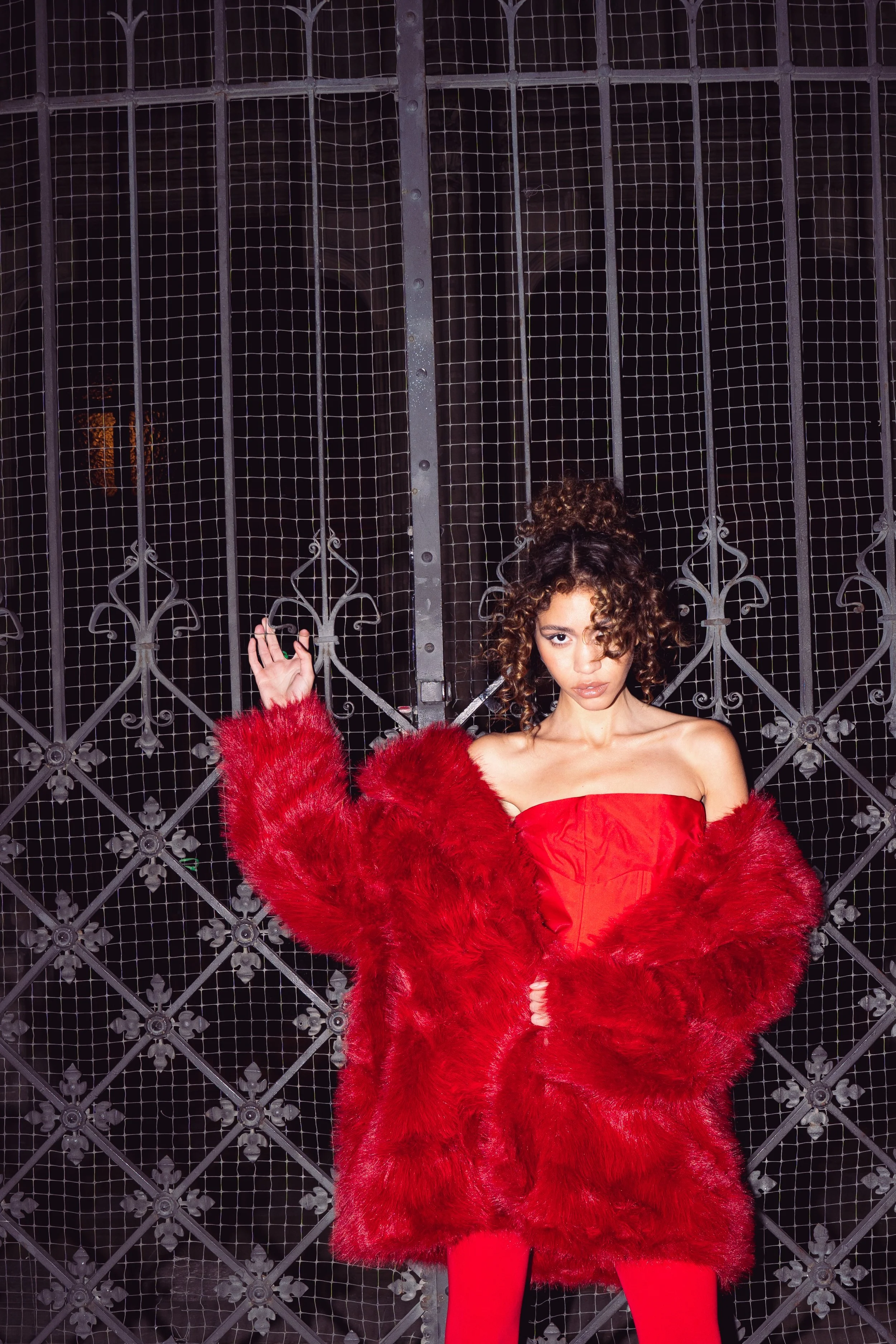 A young woman with curly hair wearing a red strapless dress and a red faux fur coat standing in front of a wrought iron gate at night.